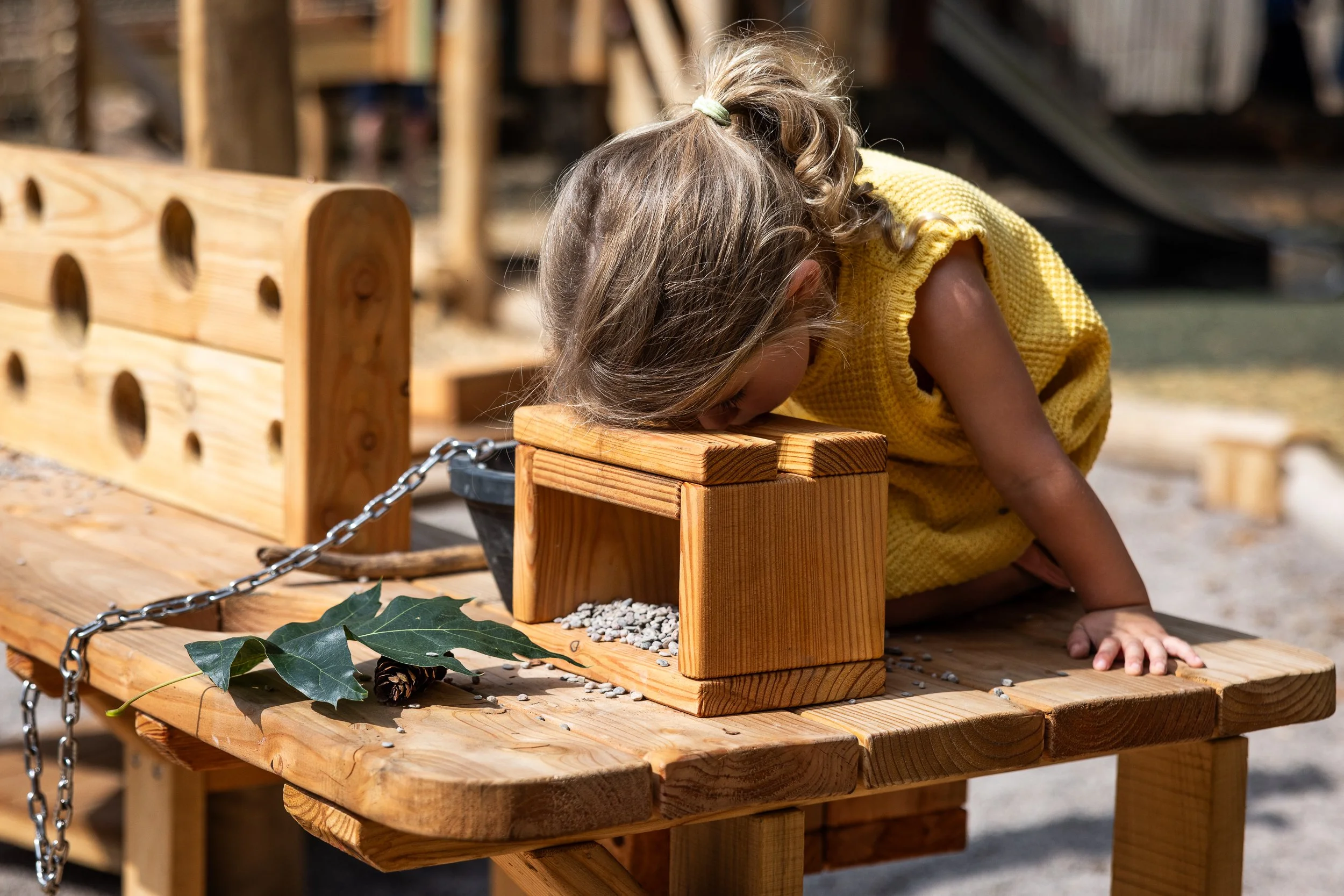 A young girl in a yellow dress with curly hair tied back, leaning her head on a wooden block on a picnic table outdoors, with wooden playground equipment and greenery in the background.
