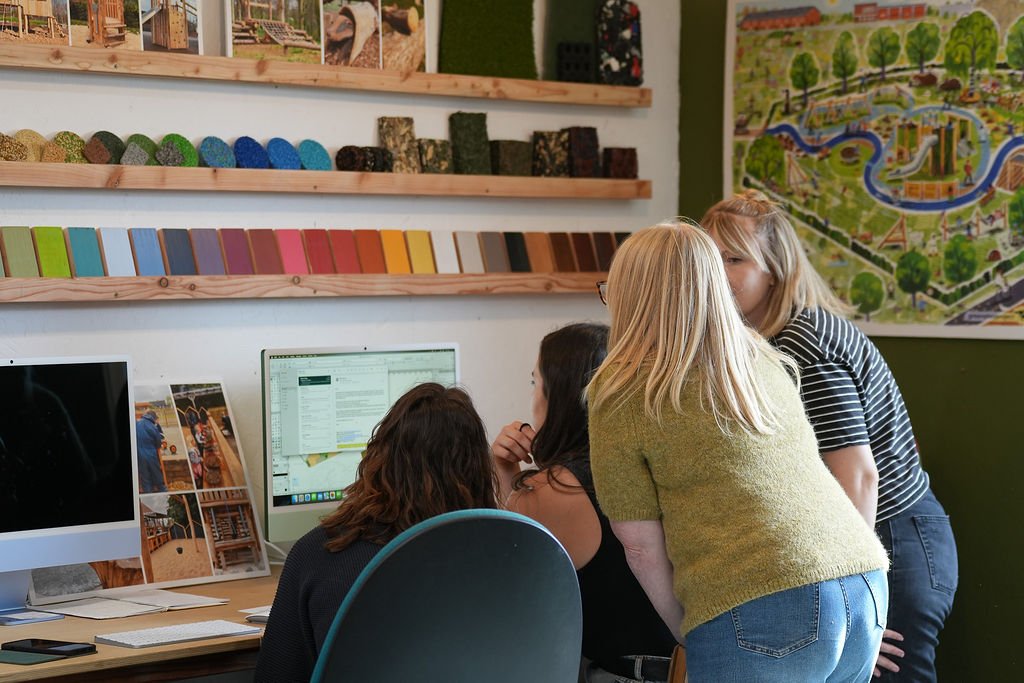 Four women working together around a computer in an office or creative workspace, with materials and a large park map on the wall behind them.
