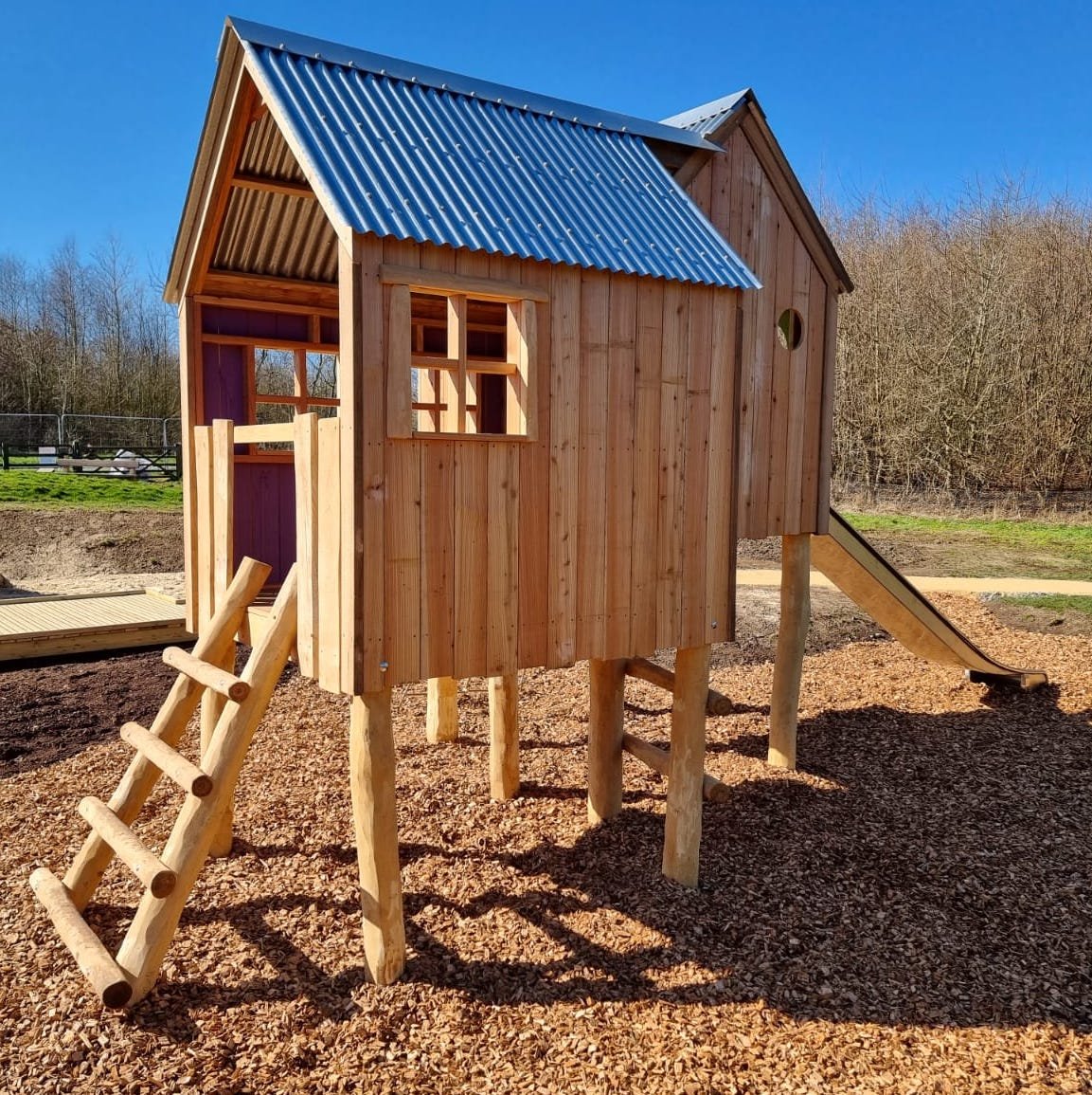 A wooden playhouse with a metal roof, raised on stilts with a small set of wooden stairs, located outdoors on a bed of wood chips.