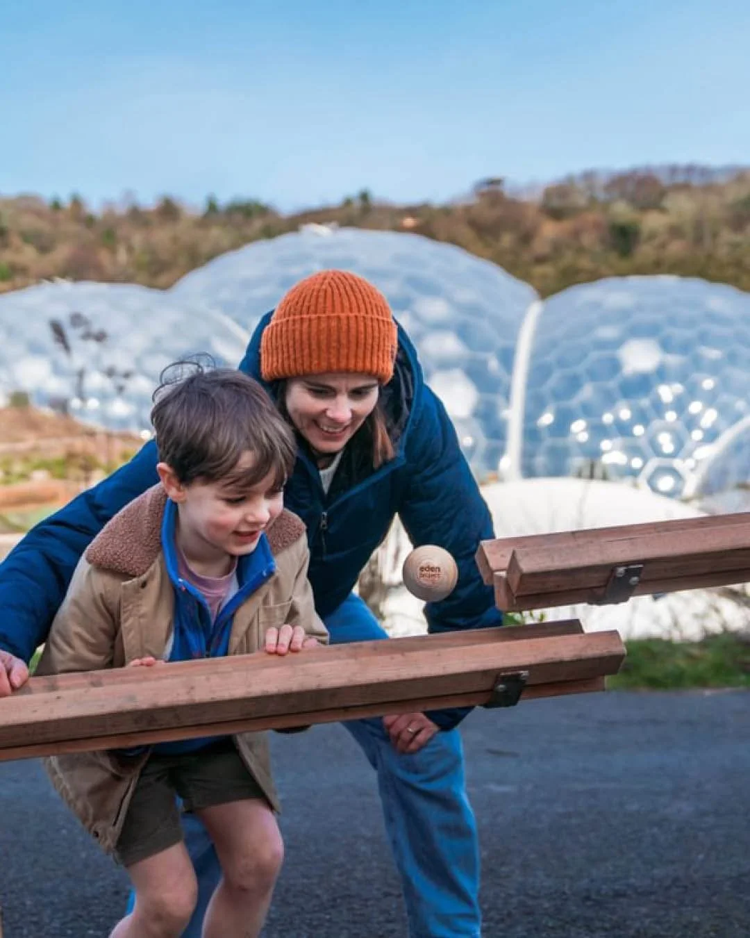 Child and adult playing on a wooden outdoor play structure designed to resemble an owl, set in a forested area with tall trees and woodland environment.