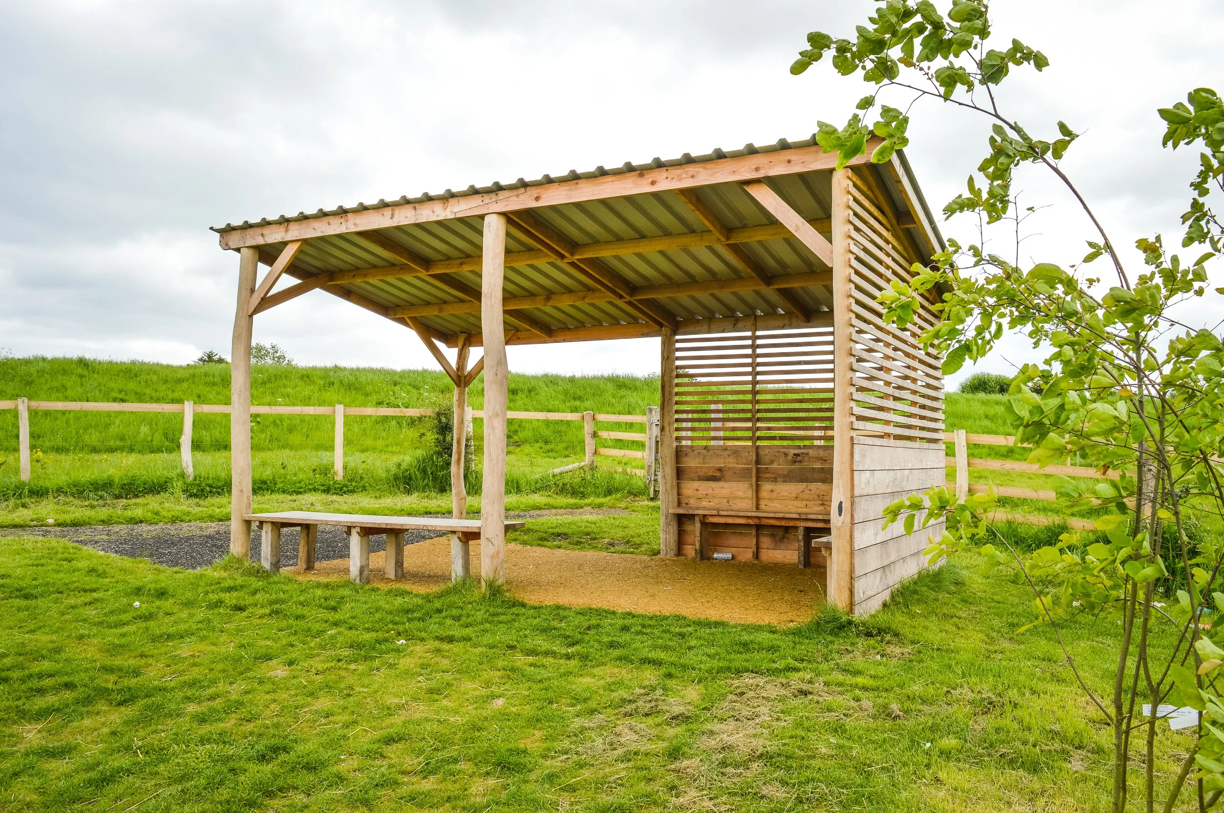 A small wooden shelter with a corrugated metal roof and partially open sides, situated in a grassy field with a fence and green hills in the background.