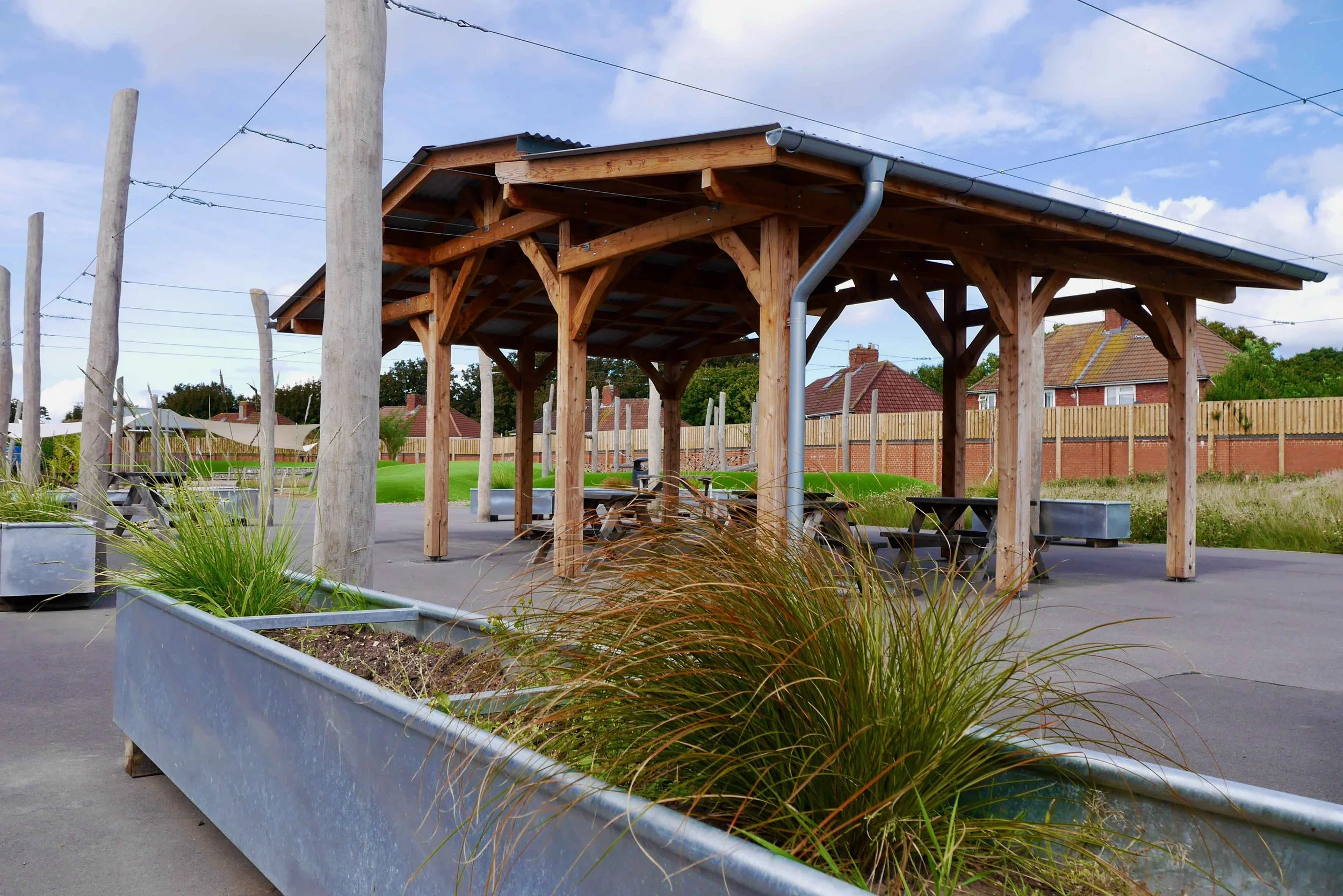 Wooden park shelter with benches, planters with grasses, and a paved area, in a neighborhood setting with houses and a wooden fence in the background.