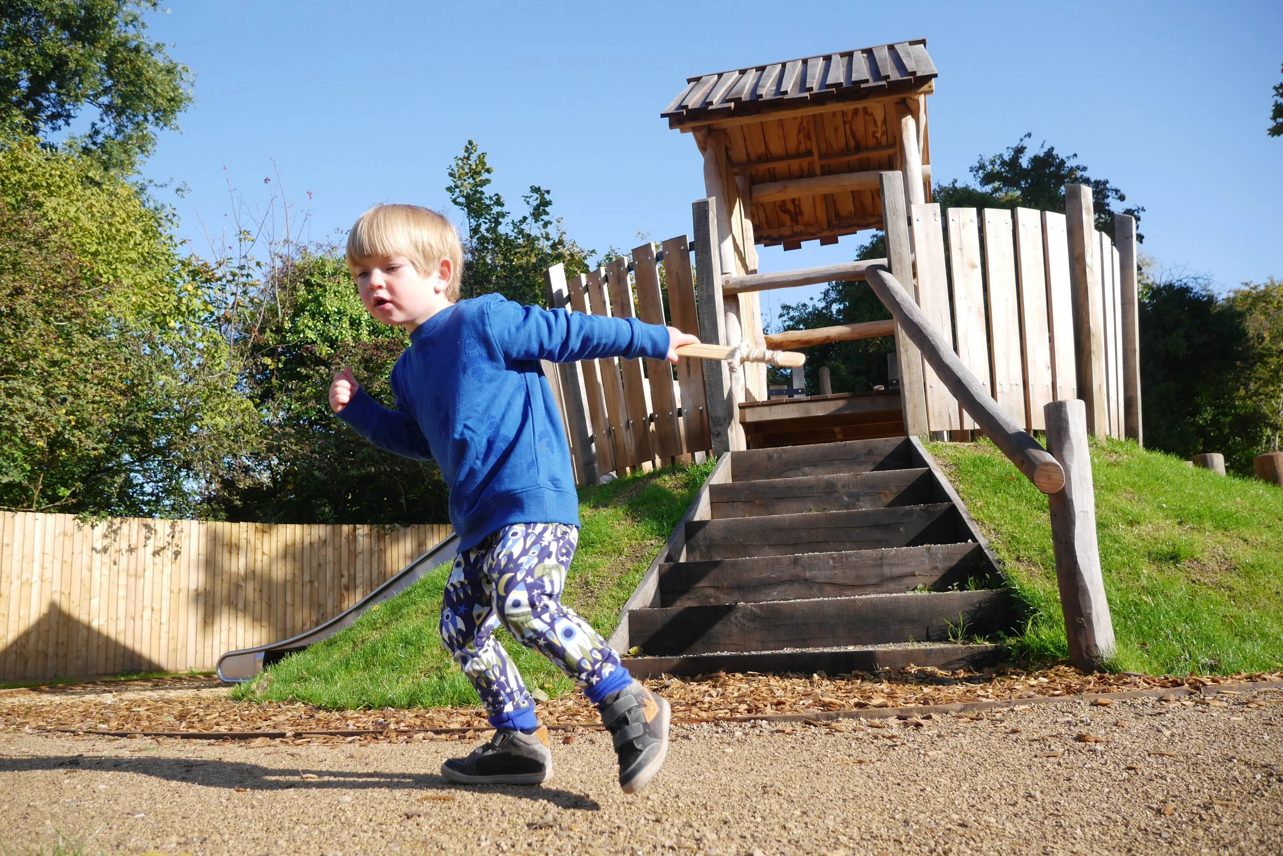 A young boy running in a playground area with a wooden play structure and slide, surrounded by trees and a wooden fence, under a clear blue sky.