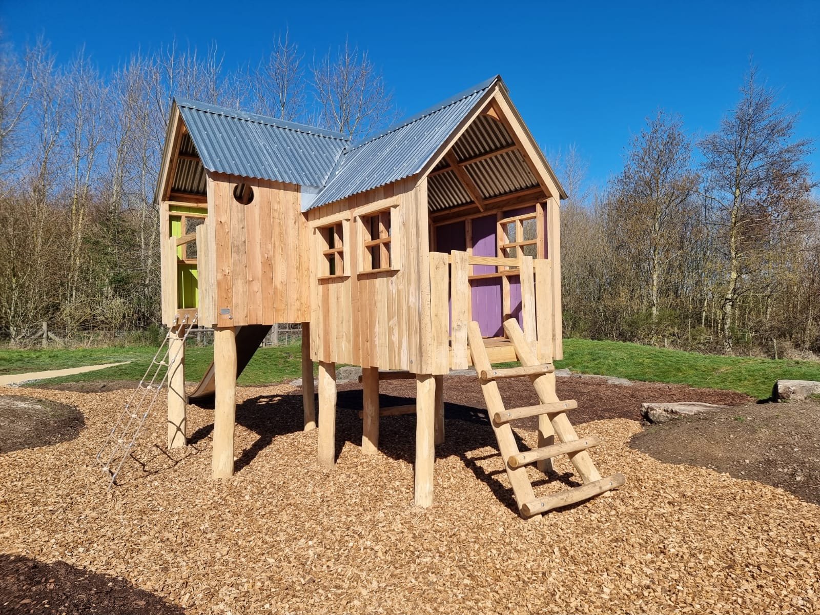 A wooden playhouse with a metal roof and small windows, elevated on stilts with a set of steps, in an outdoor park with trees in the background.