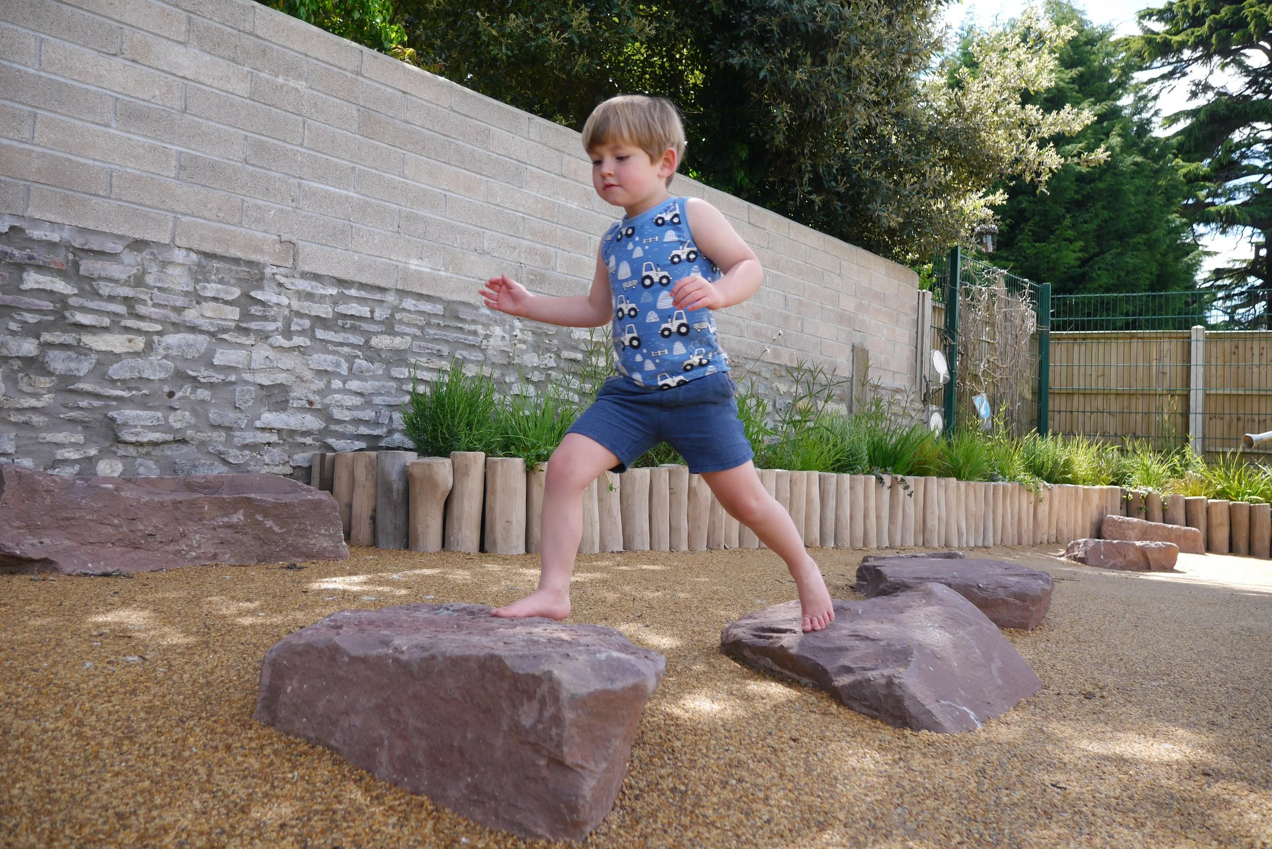 A young boy in a blue tank top with car patterns and blue shorts balancing on large rocks in a backyard garden area with a brick wall, green plants, and a fence in the background.