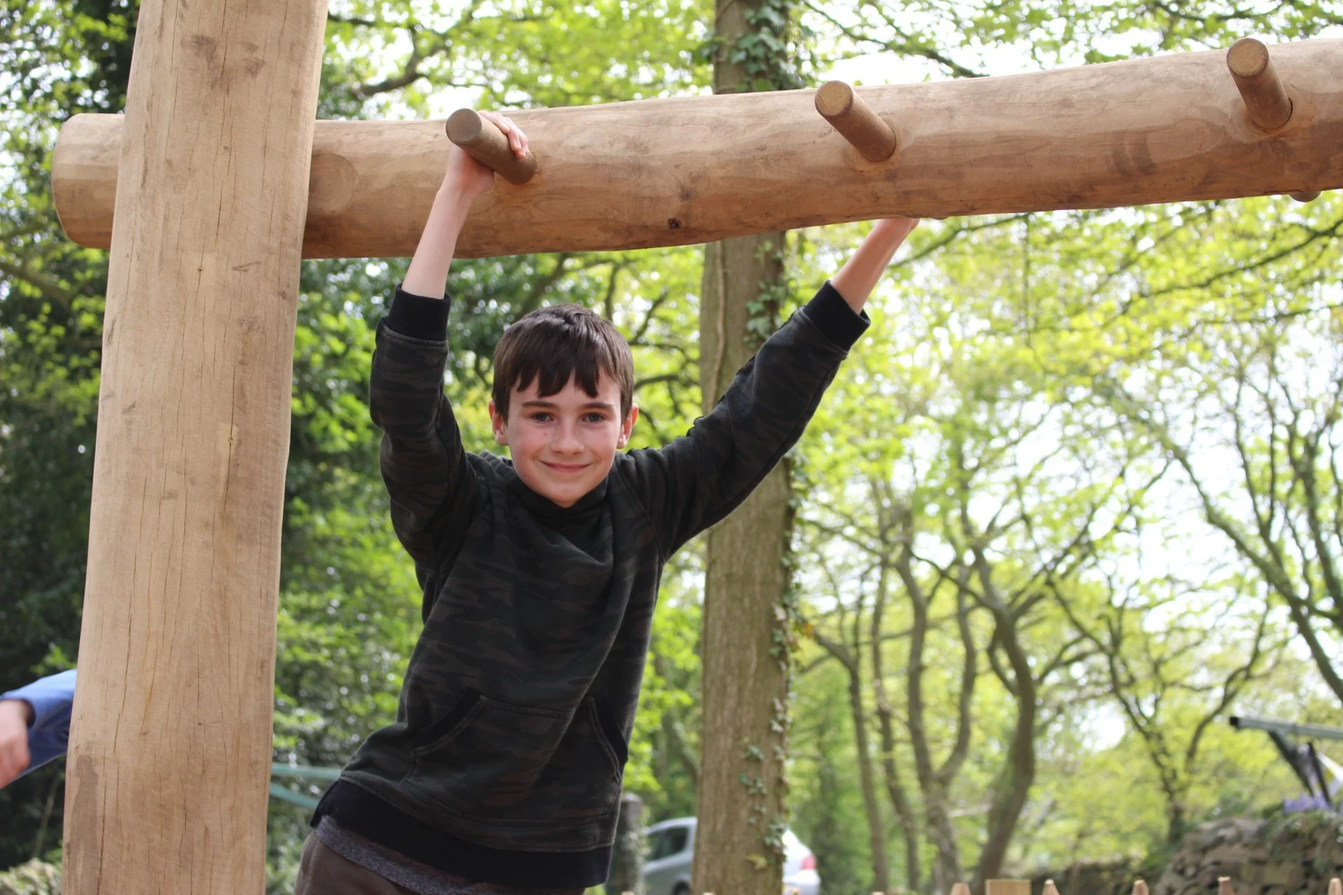 Young boy hanging from a wooden outdoor playground structure in a park with green trees in the background.