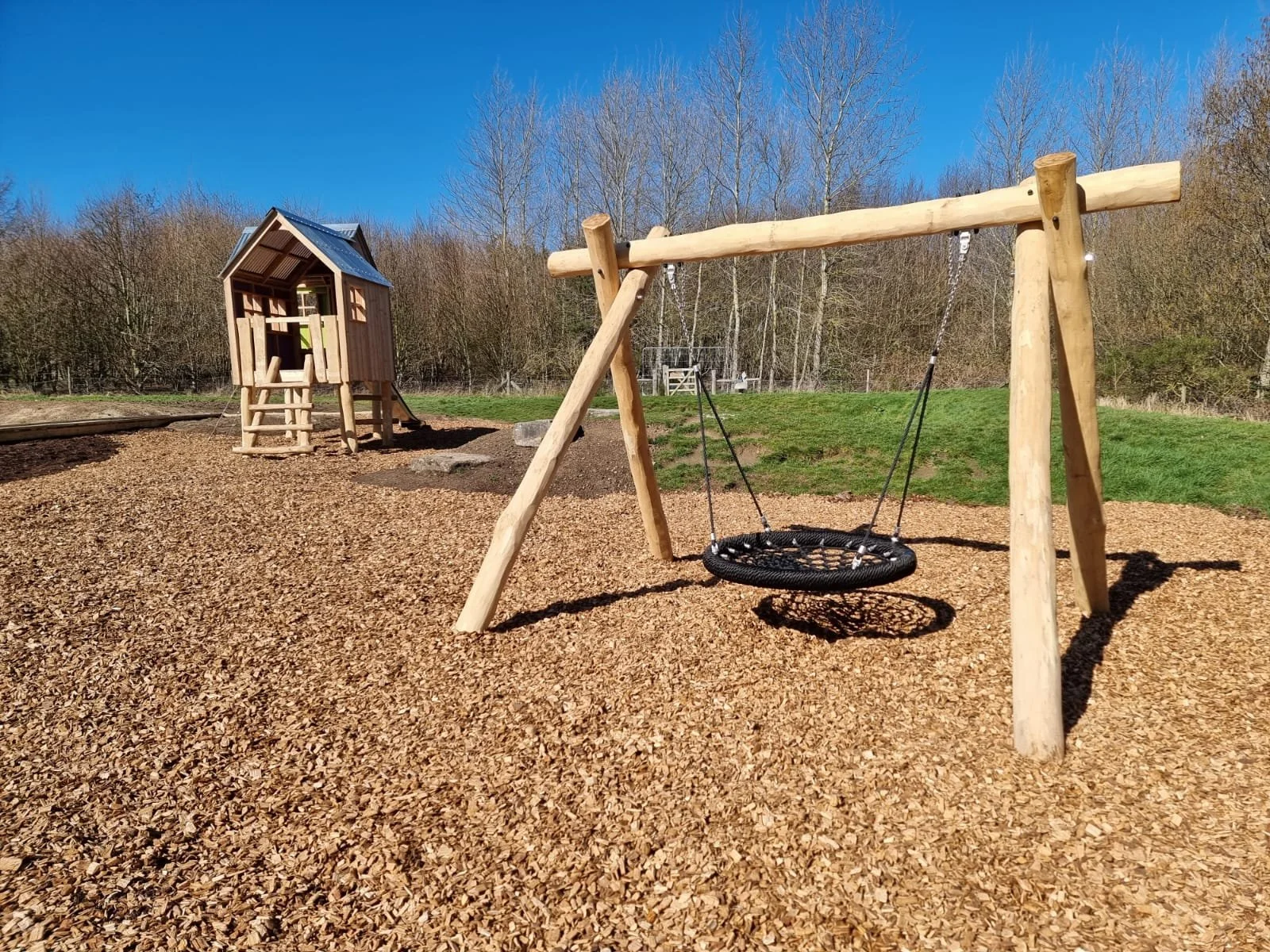 A playground with a wooden swing set and a wooden playhouse on a bed of wood chips on a sunny day with clear blue sky.