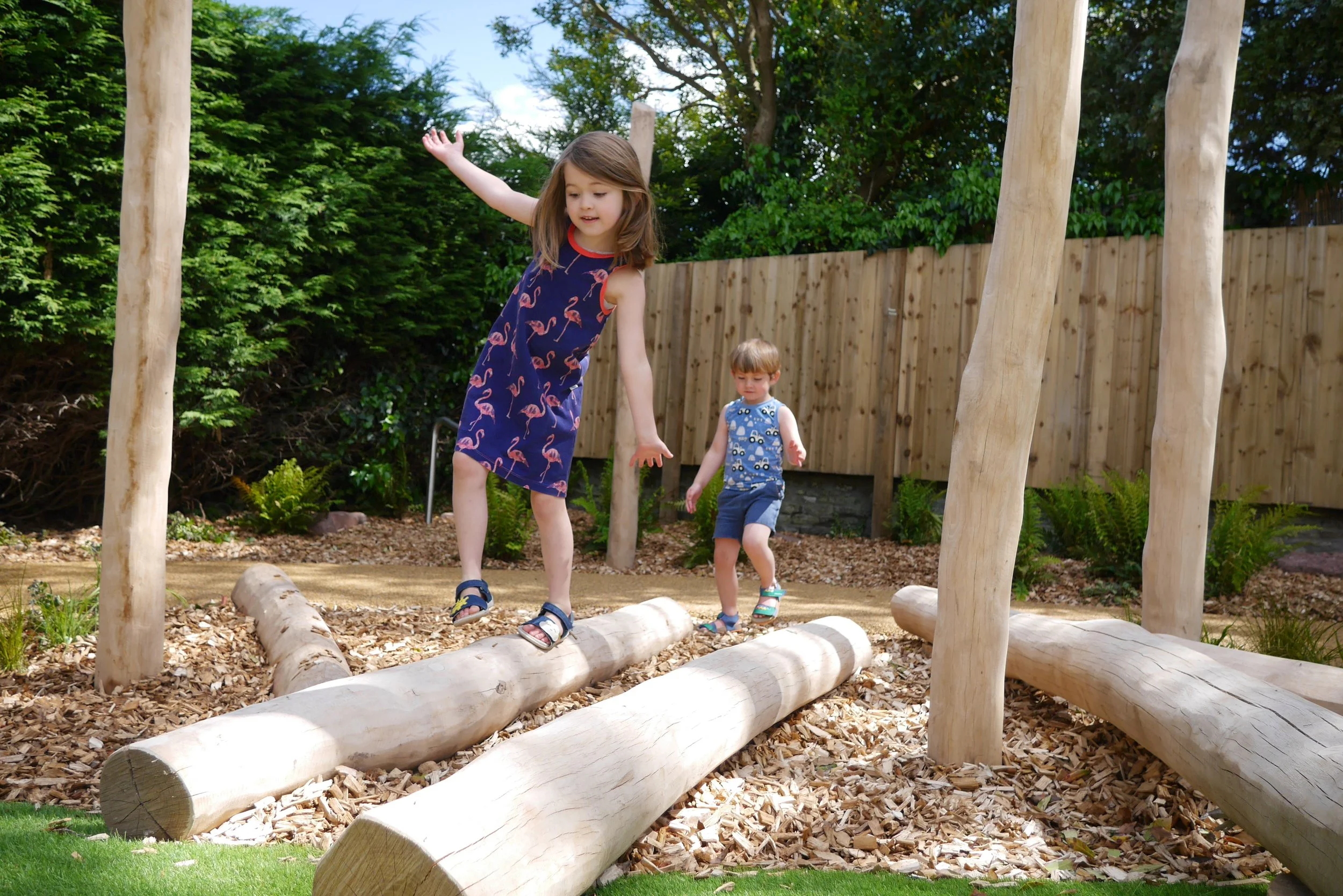 Two children playing on wooden logs in a backyard or playground area with greenery and a fence in the background.