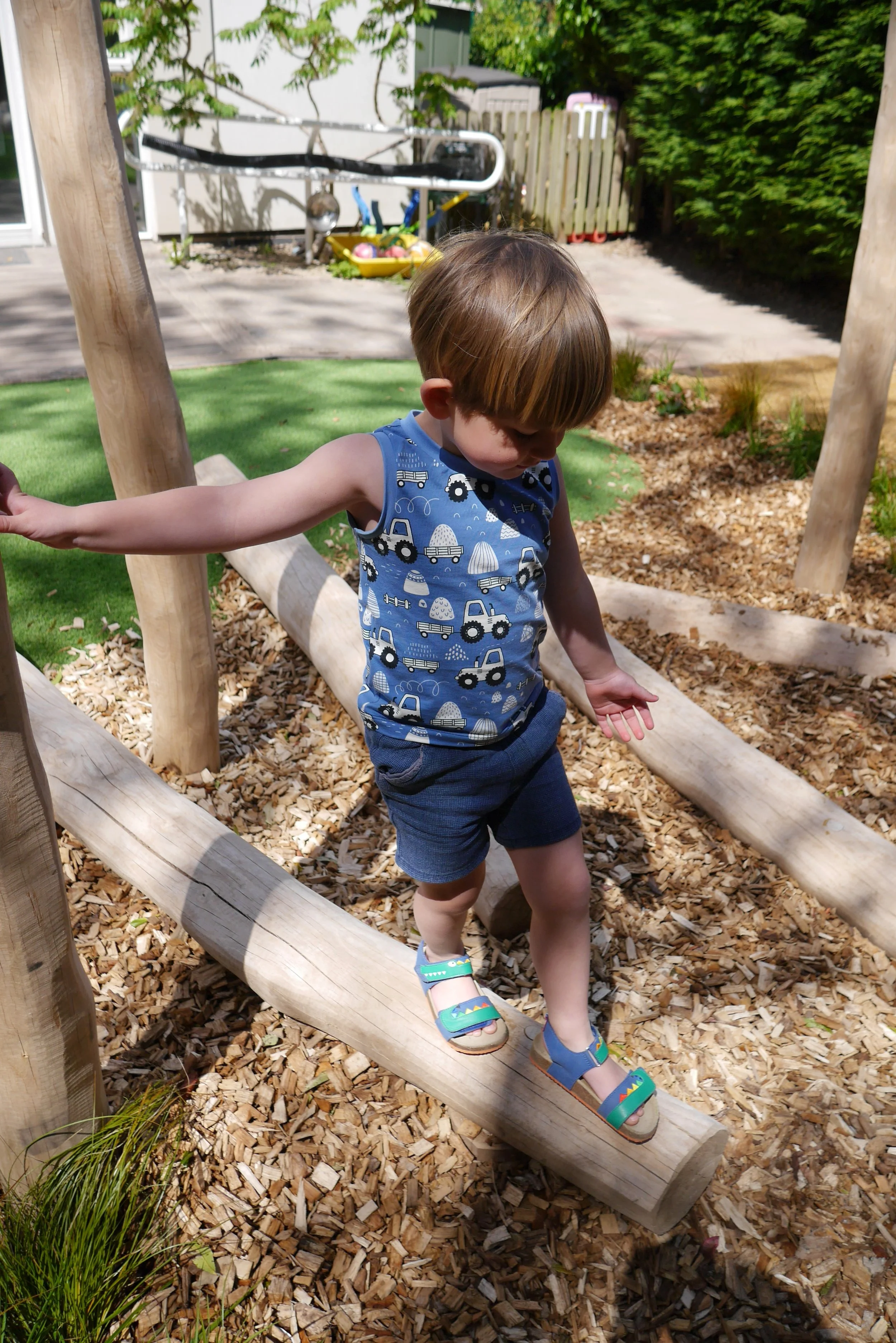 A young boy with brown hair, wearing a blue tank top with tractor and construction vehicle prints, blue shorts, and colorful sandals, balancing on a wooden log in a backyard playground during daytime.