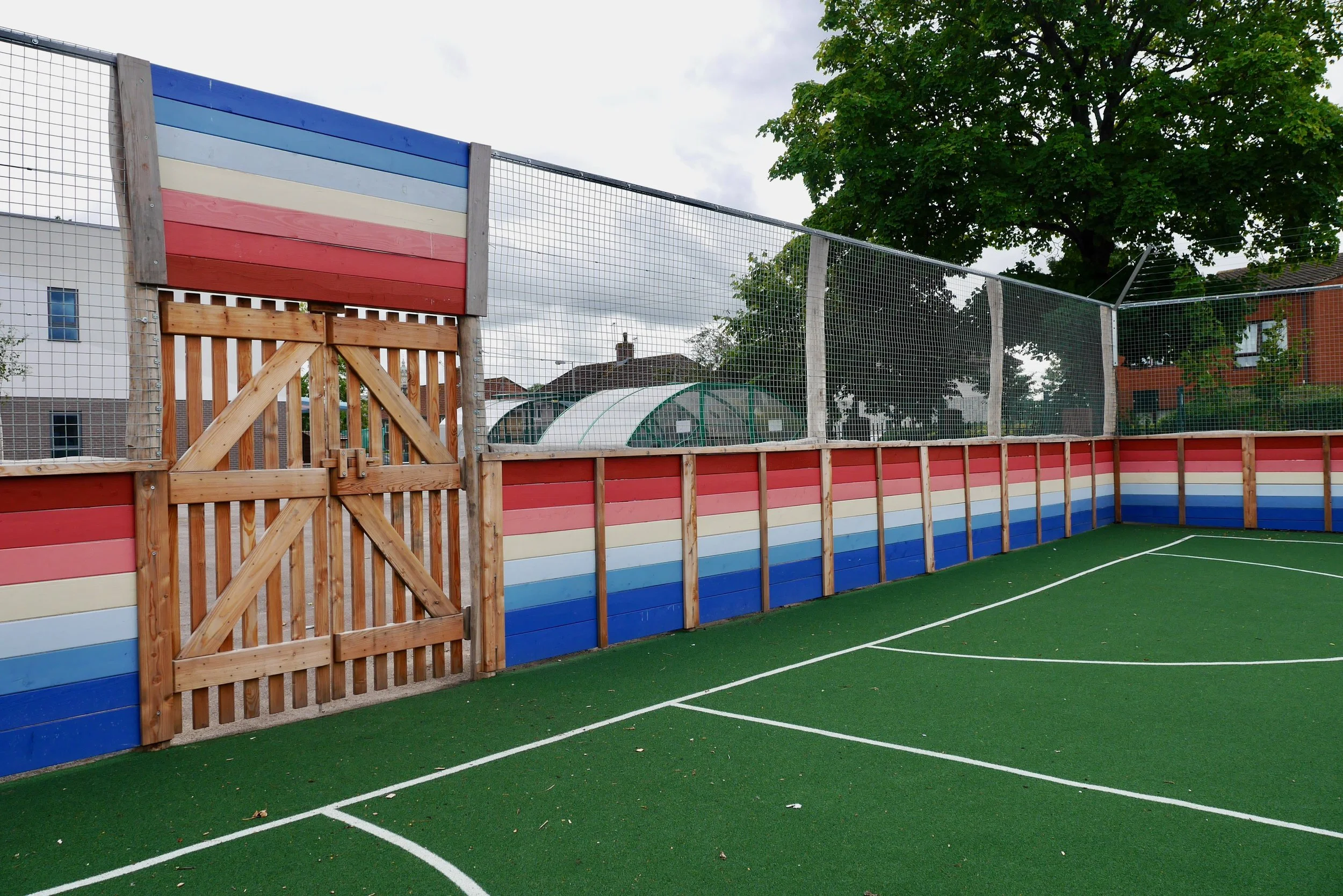 A small enclosed basketball court with a green artificial turf surface and a colorful wood-paneled fence with red, white, and blue stripes. There is a wooden gate in the fence and a large tree in the background.