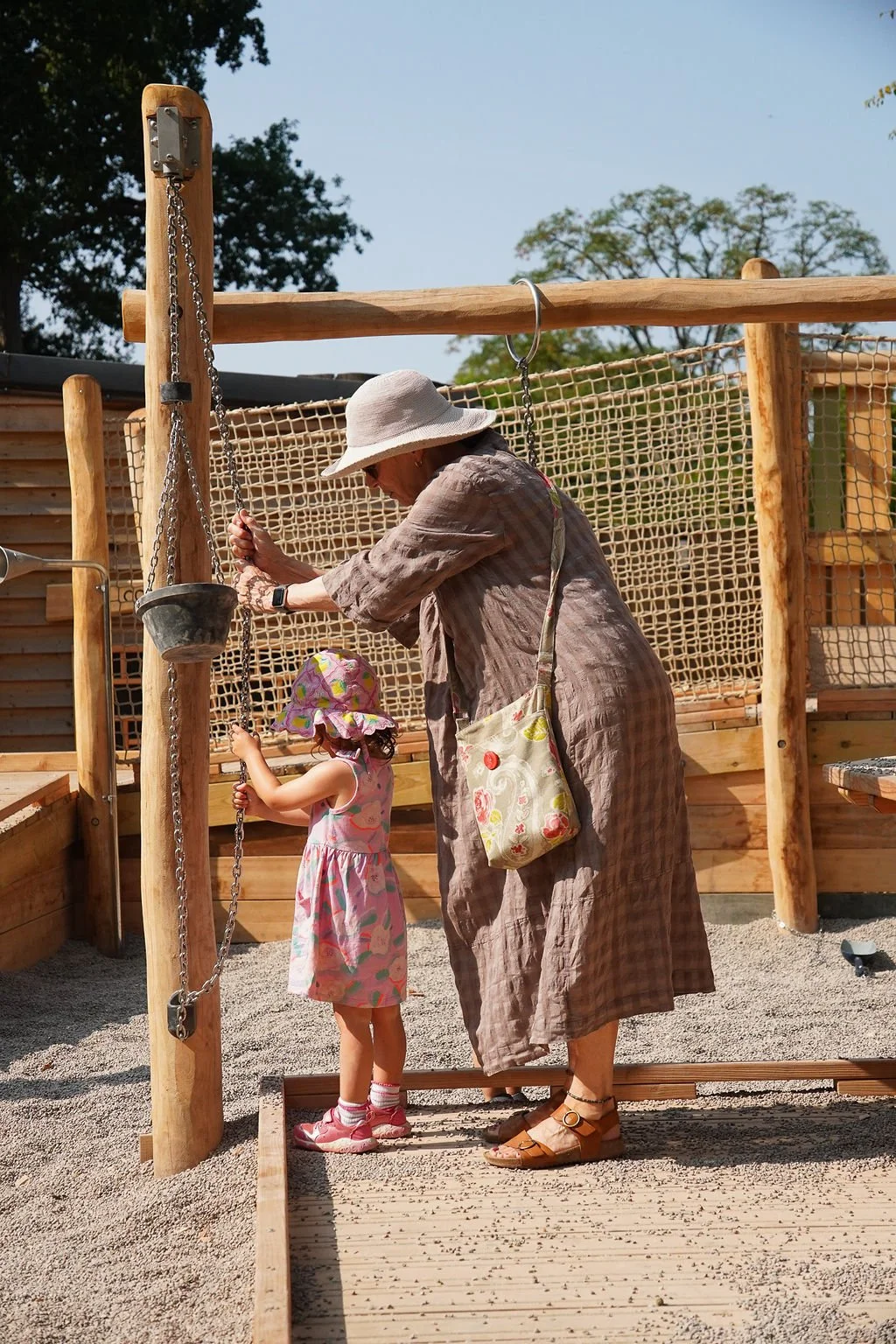 A woman and a young girl at a playground swing set, with the woman adjusting the swing and the girl holding onto the chains, both wearing summer hats.