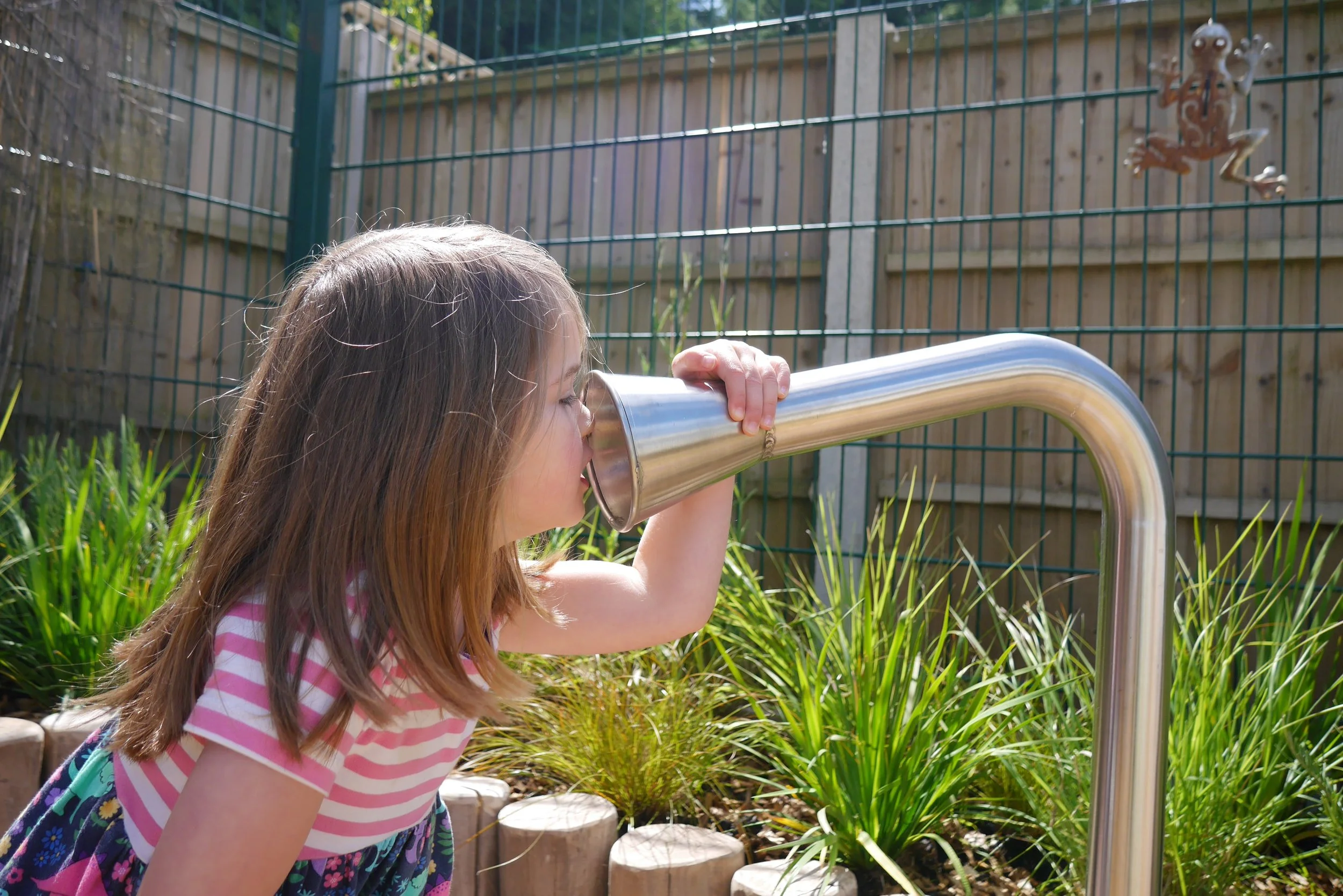 Young girl with brown hair wearing a pink and white striped shirt, looking through a metal telescope in a garden with wooden borders and green plants.