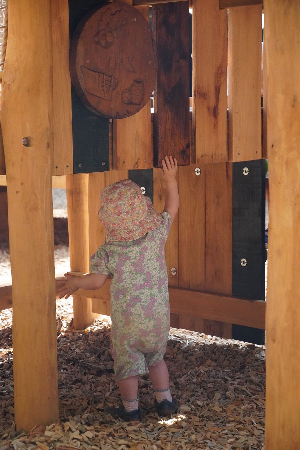A young child wearing a floral dress and sun hat plays under a wooden play structure outdoors, reaching up to touch the wood panel with a carved oak tree and acorn.