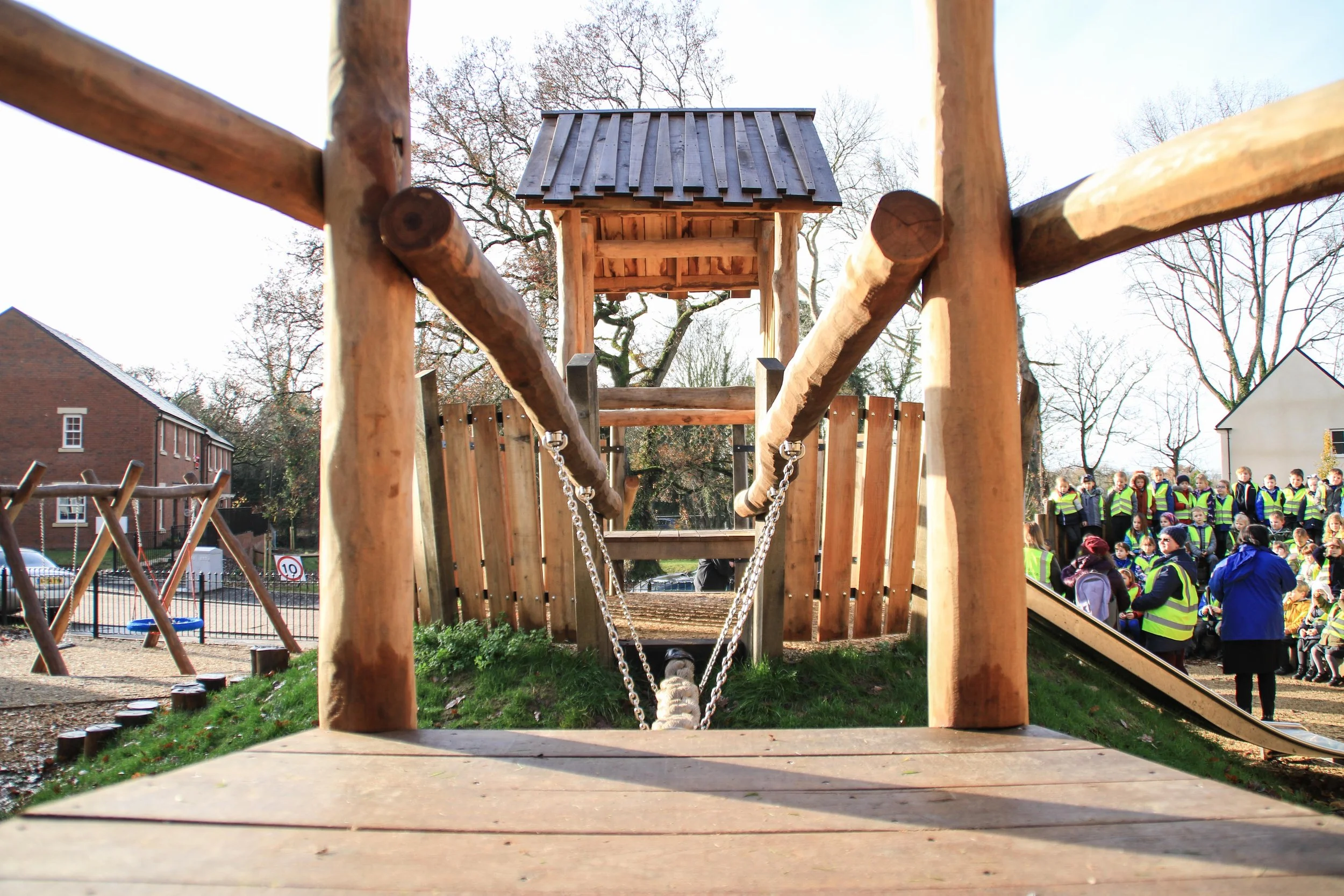 Photo taken from the ground level of a wooden playground structure with a bridge and a small shelter on top. In the background, children wearing yellow safety vests are gathered, and there are trees and buildings around.