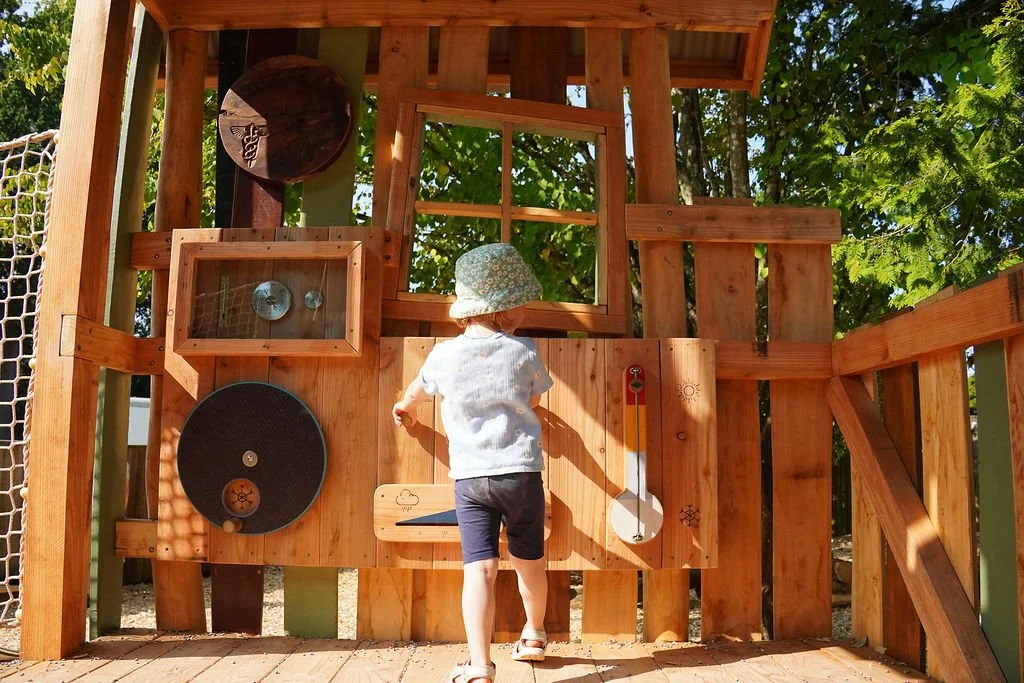 Child wearing a hat and shorts playing at a wooden outdoor sensory wall with various textured and interactive elements.