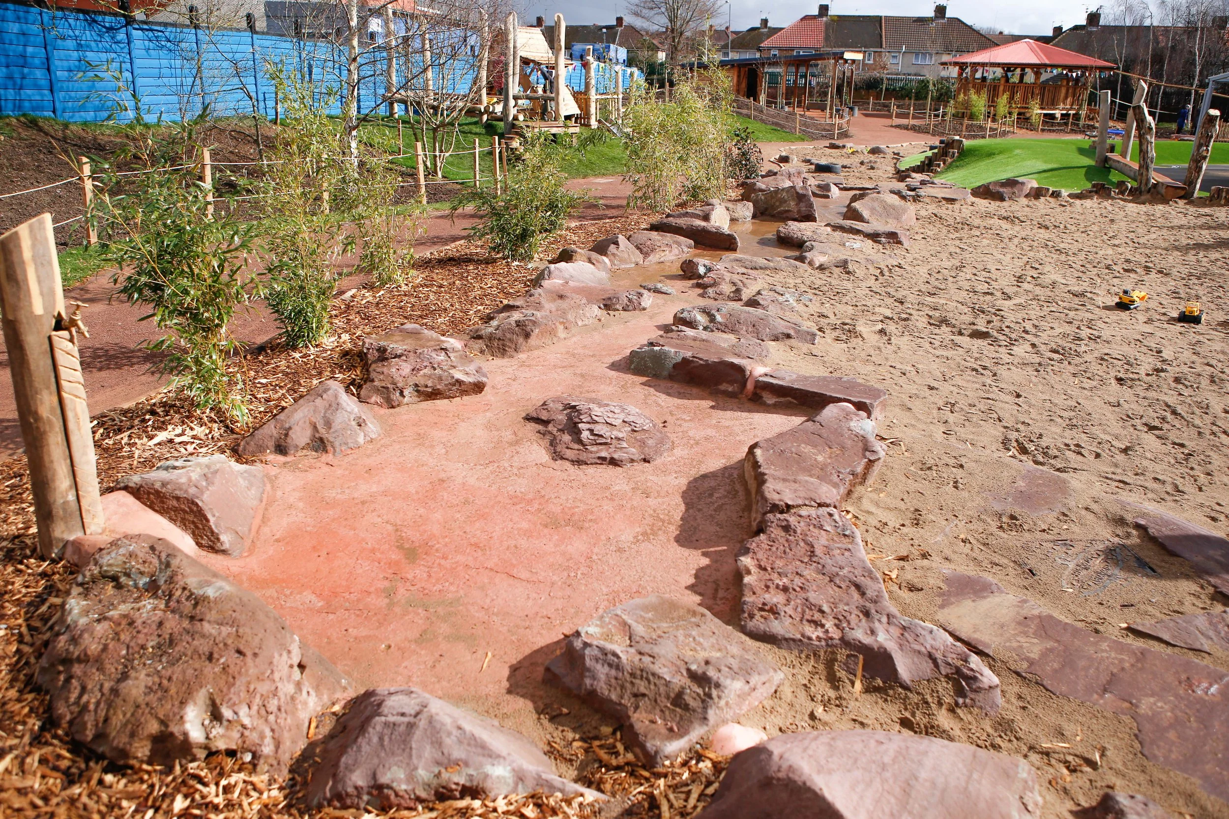 An outdoor playground area featuring a sandy surface with a border of large reddish-brown rocks. There are plants along the edge of the play area, and in the background, there are additional play structures, a small gazebo, and residential houses. To