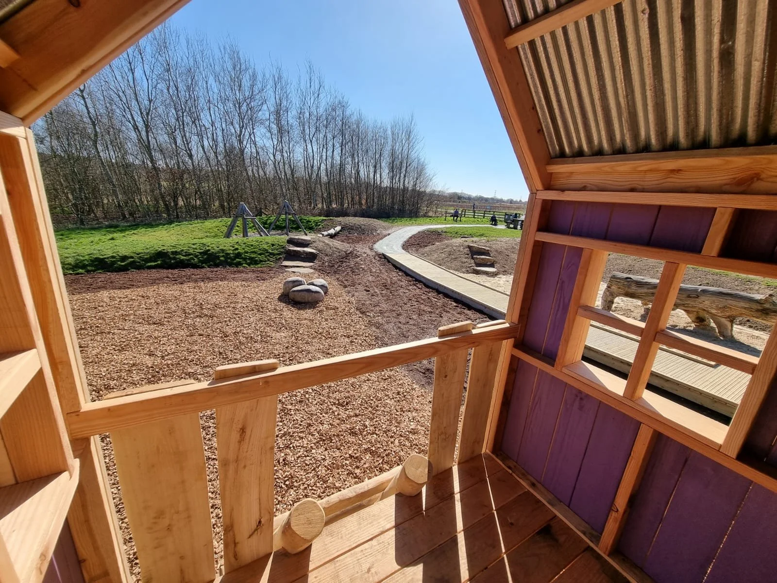 View from inside a small wooden playhouse looking out onto a playground with a gravel path, a swing set, green grass, trees, and children playing in the distance on a sunny day.