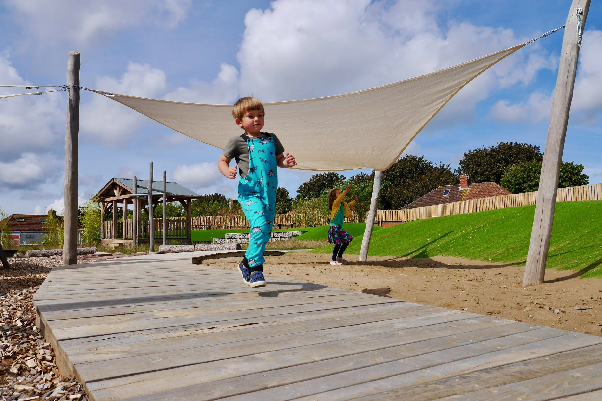 Children playing in a park with a wooden bridge, sandy ground, green grassy hill, and a beige shade cloth overhead, under a partly cloudy blue sky.