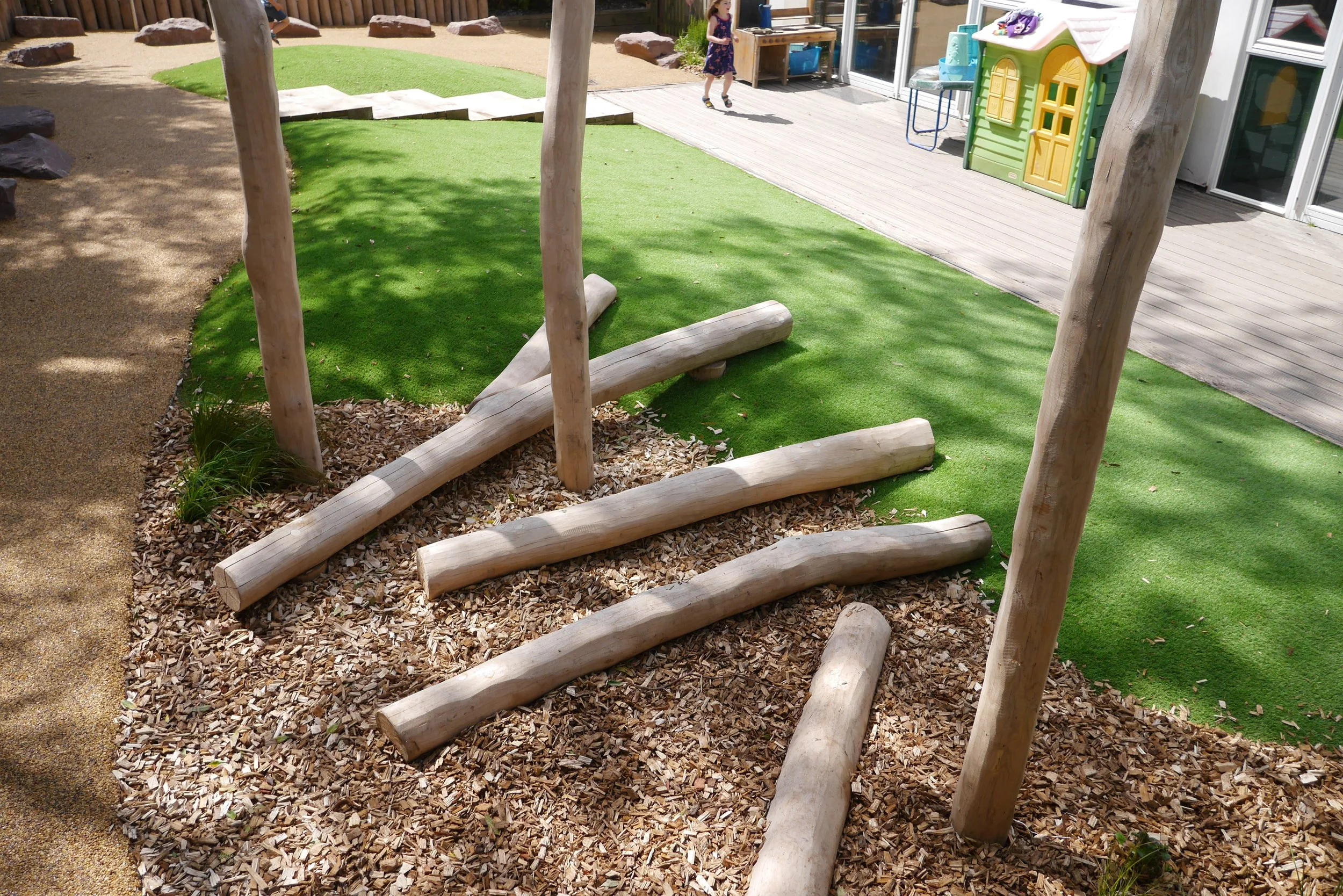 Children's playground with wooden beams, artificial grass, and a playhouse in the background.