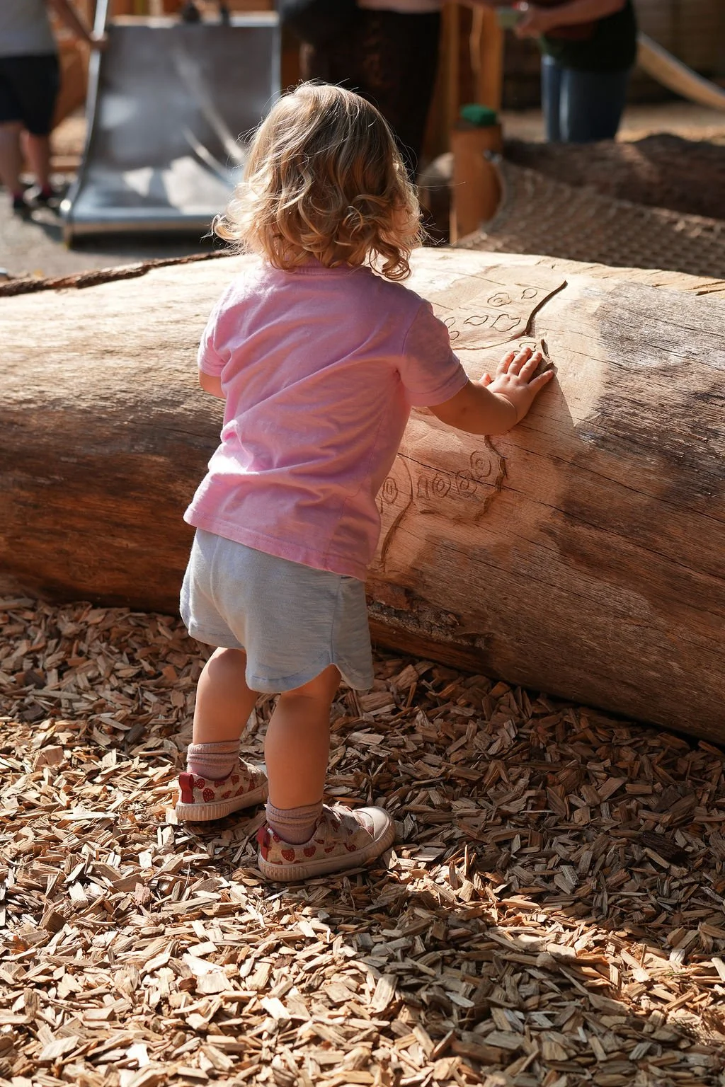 A young girl with curly blonde hair, wearing a pink T-shirt, grey shorts, and patterned sneakers, is standing outdoors on wood chips, touching and examining a large wooden log with carved drawings on it.