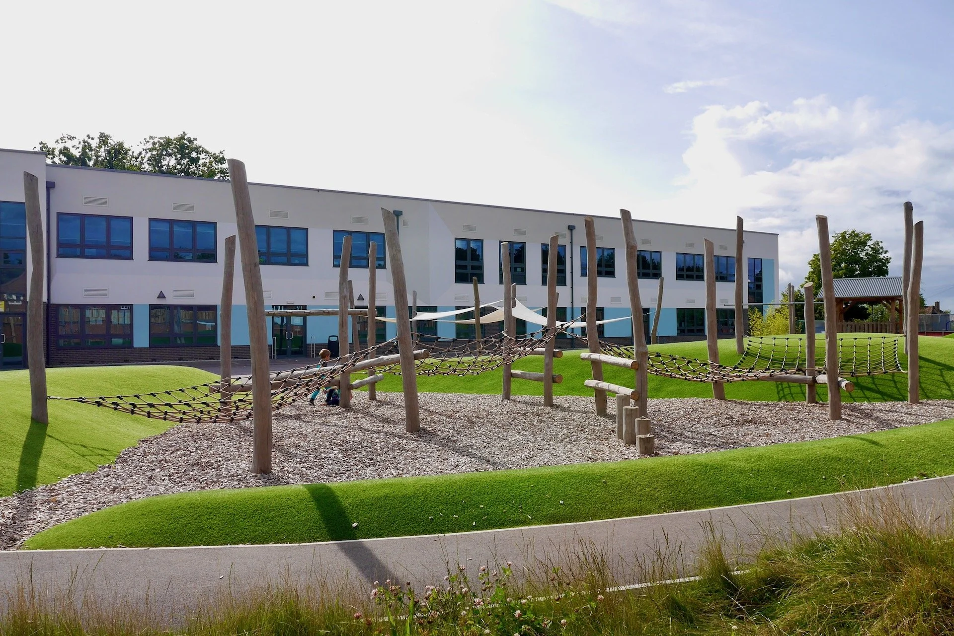 Modern school building with large blue-tinted windows and a well-maintained grassy playground with wooden play structures and climbing nets. Bright daylight with partly cloudy sky.