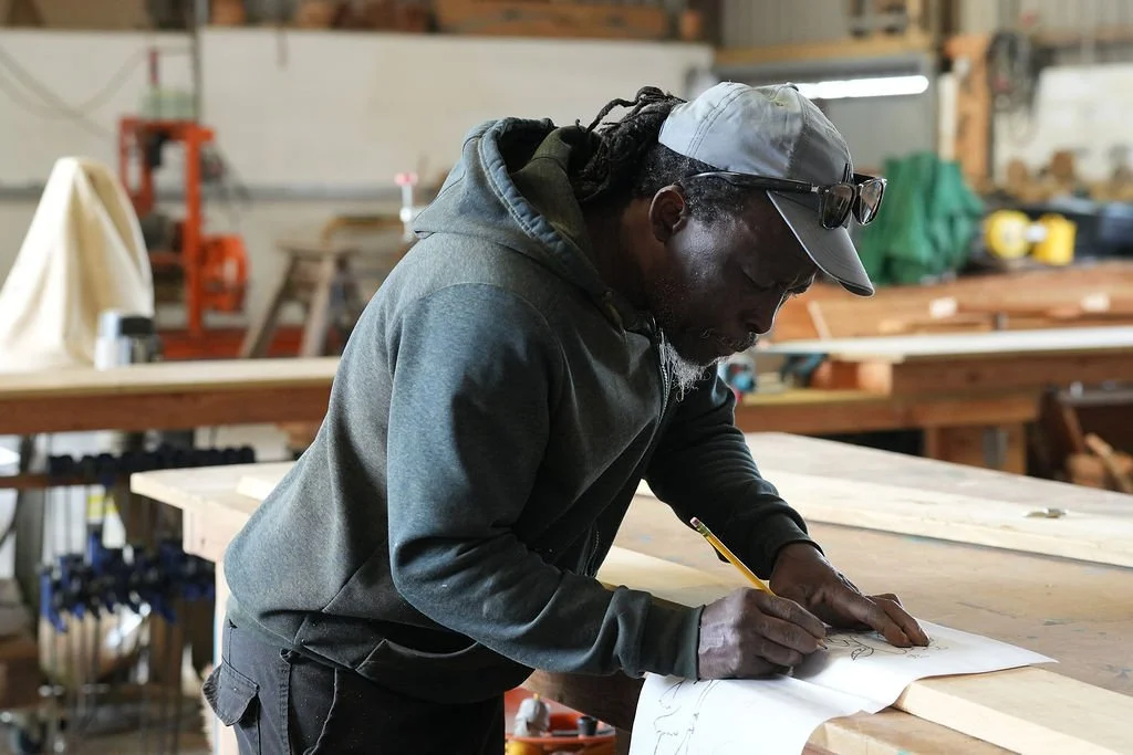 A man wearing a gray hoodie, gray cap, and glasses on his cap, works on a large piece of paper at a woodworking workshop.