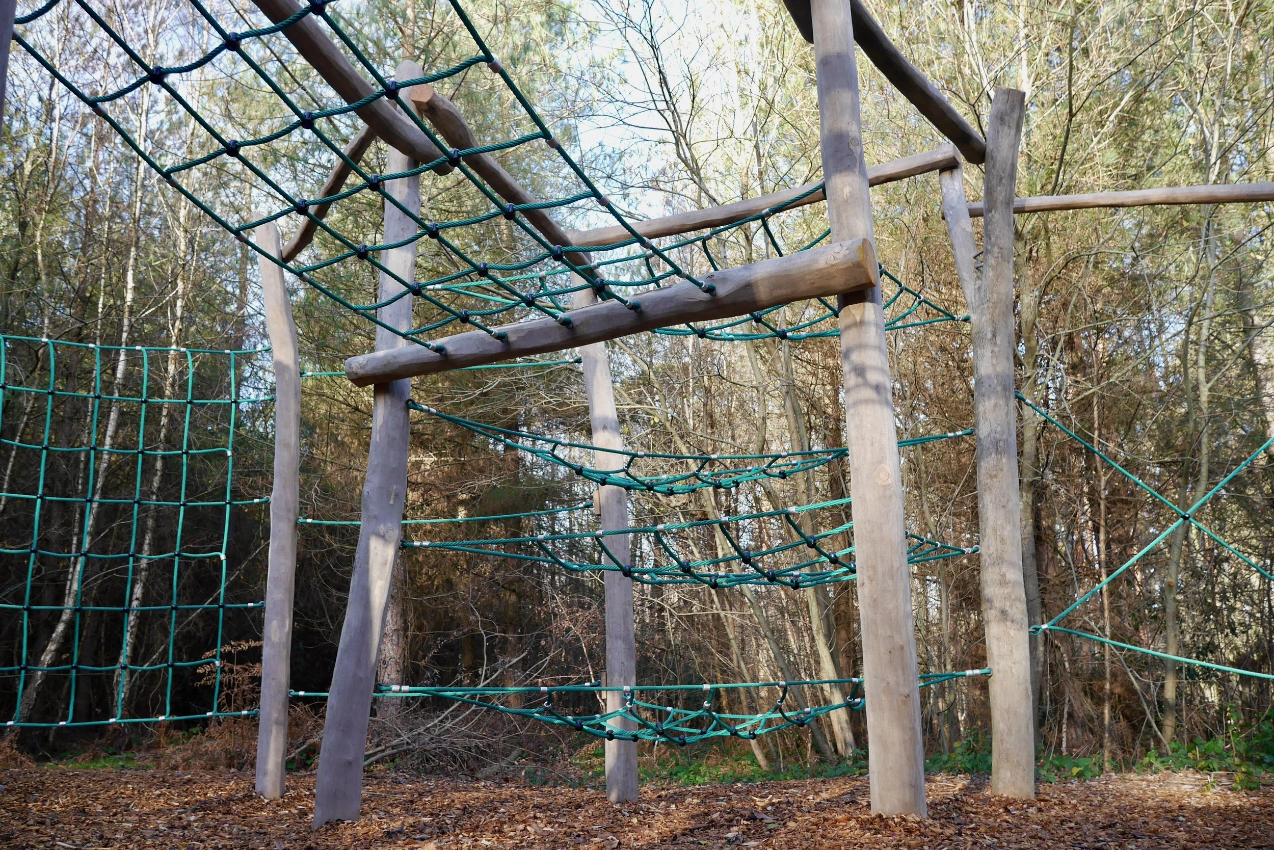 Climbing structure in a wooded outdoor play area, with wooden poles and green ropes forming platforms and netting for children to climb.