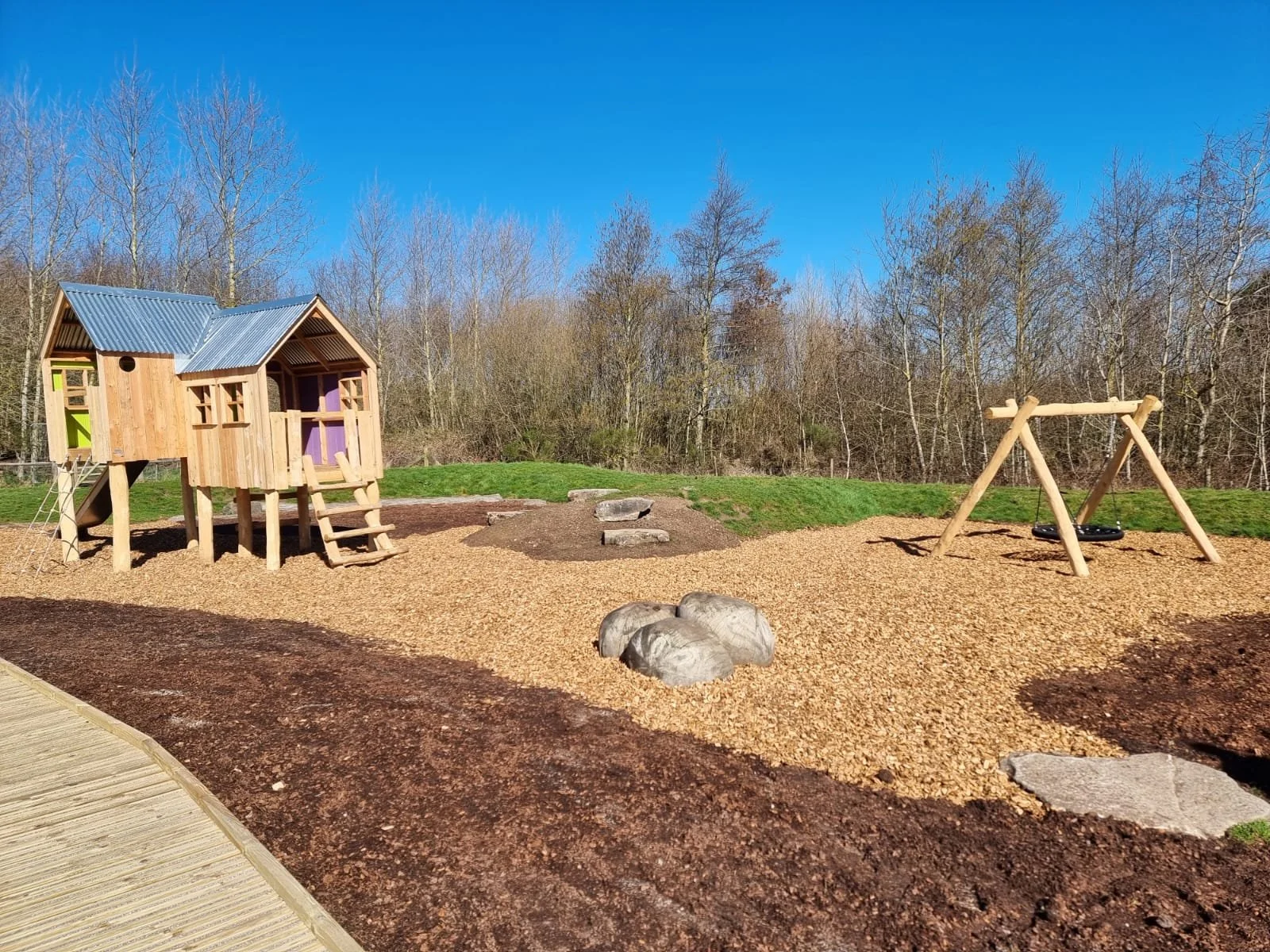 Children's playground with a wooden playhouse on stilts, a sandbox area with rocks, and a wooden swing set, set on a bed of mulch, with trees in the background under a clear blue sky.