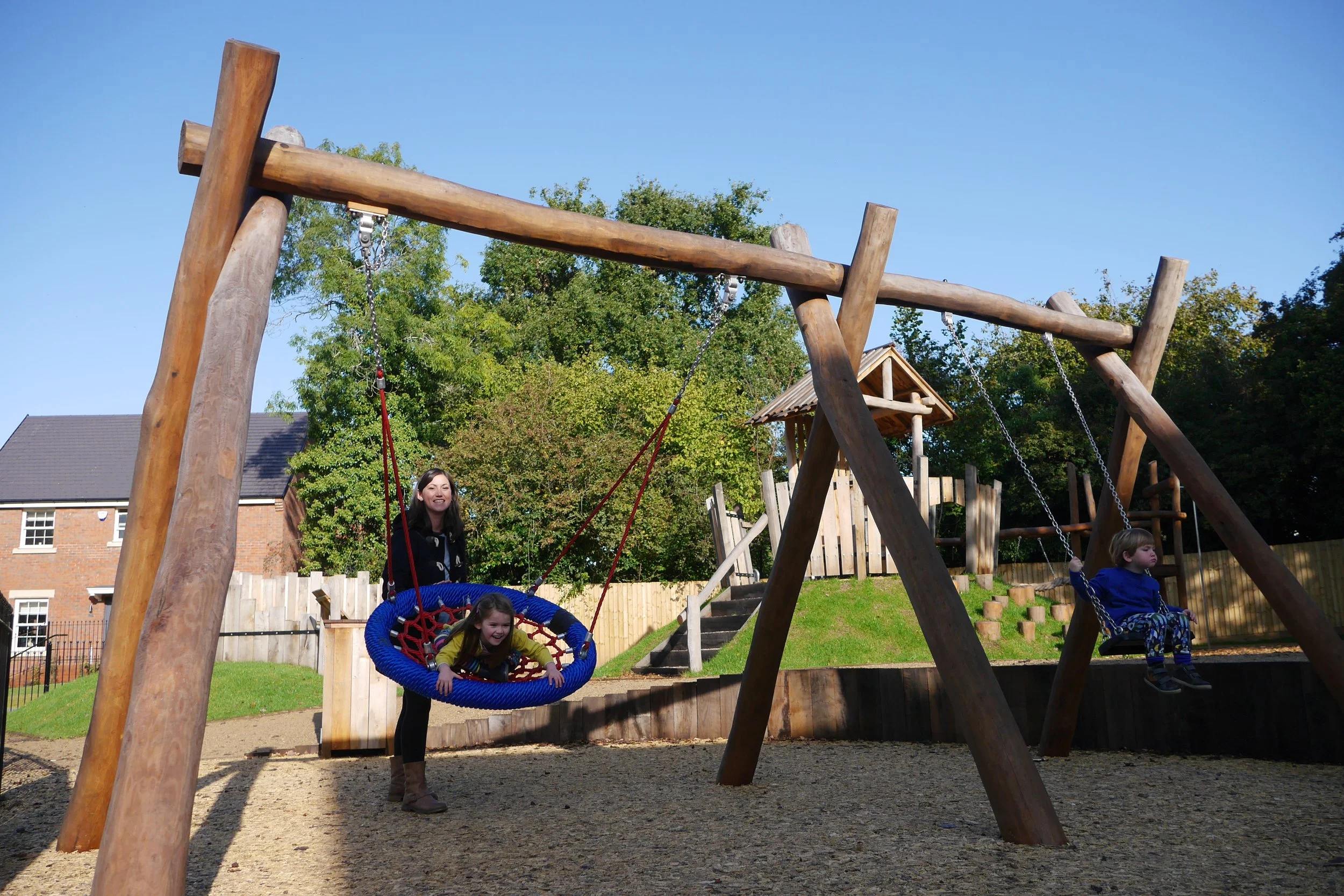 Children playing on a wooden playground with swings, with a woman standing nearby. There are trees and houses in the background.