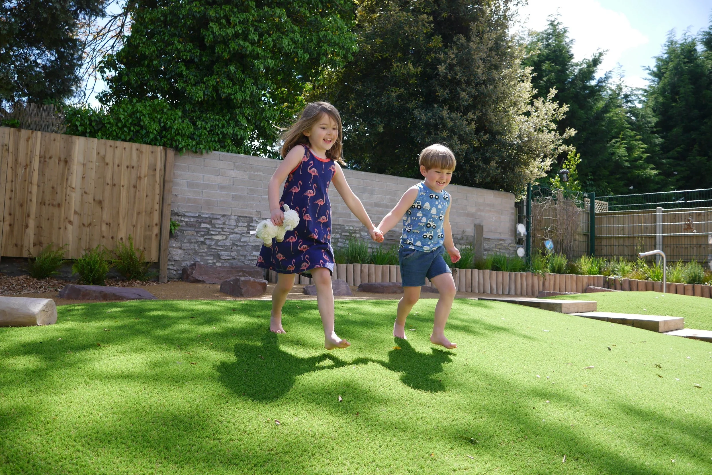 Two children, a girl and a boy, happily hold hands and run barefoot on a well-maintained green lawn in a backyard, with trees and a wooden fence in the background on a sunny day.
