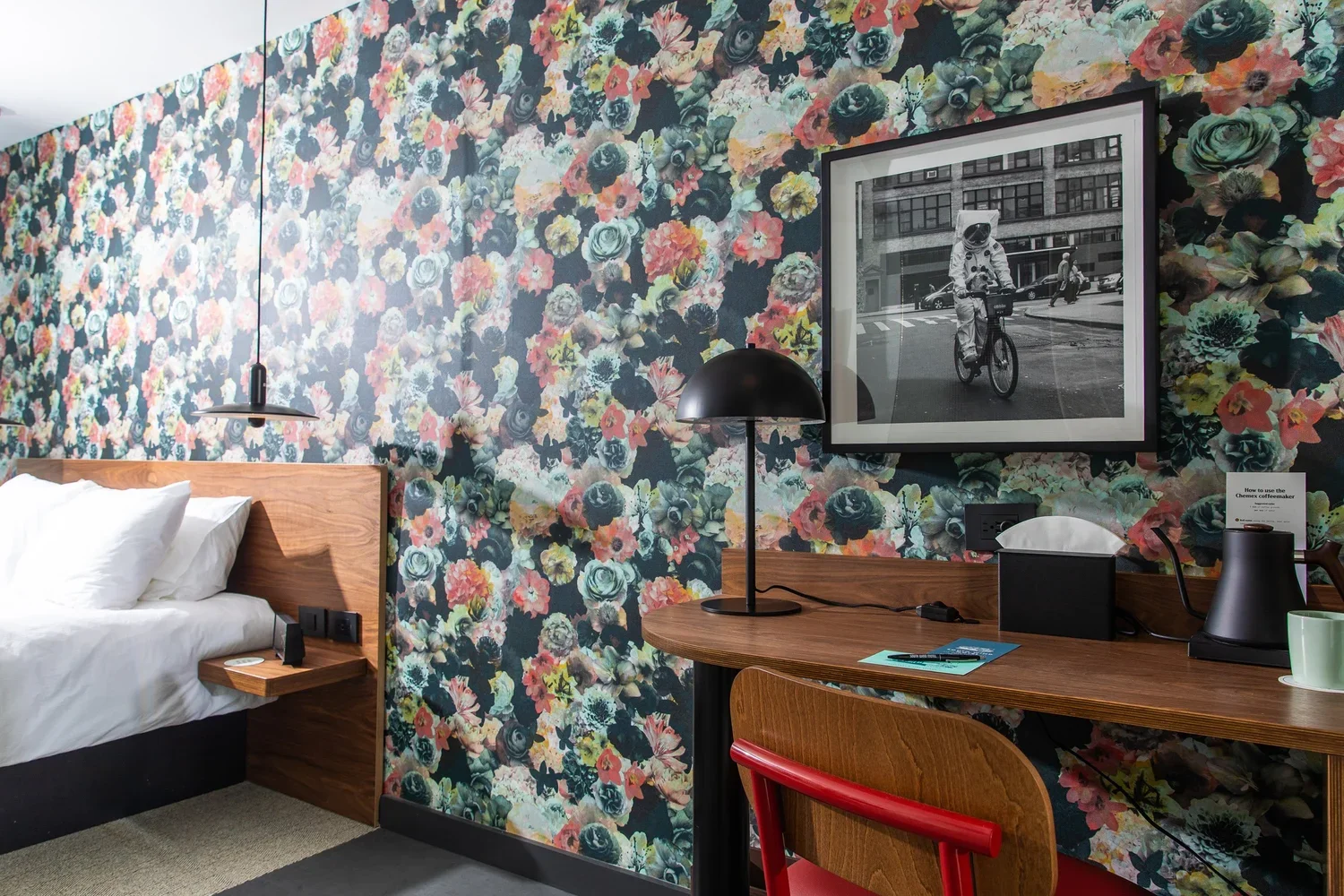 Interior of a hotel room with floral wallpaper, a bed with white linens, a wooden headboard, a nightstand with a small lamp and remote, a black wall-mounted lamp, a wooden desk with a black lamp, a framed black-and-white photo of a cyclist on a city street, and a chair with a red backrest.