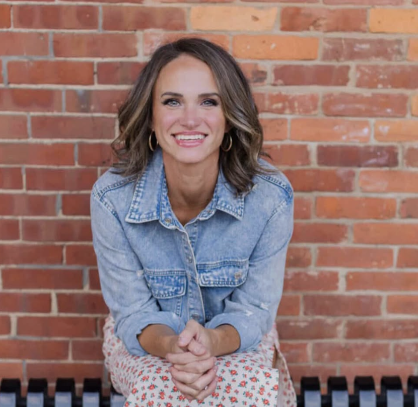 A woman with shoulder-length wavy brown hair, wearing hoop earrings and a denim jacket, sitting against a red brick wall, smiling at the camera.