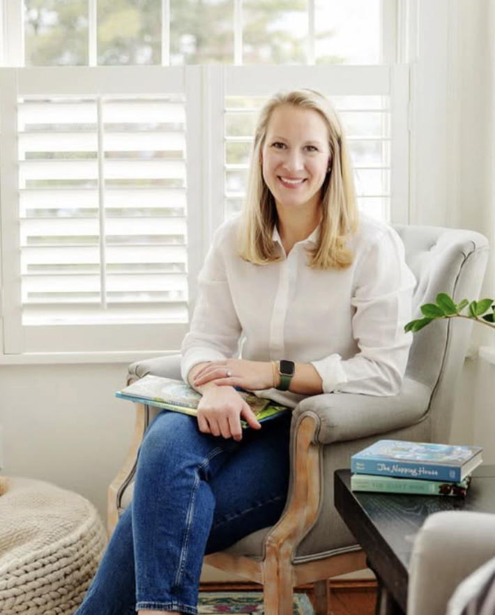 A woman with blonde hair smiling, sitting on a light-colored armchair in a room with a large window with white blinds, holding a closed book.