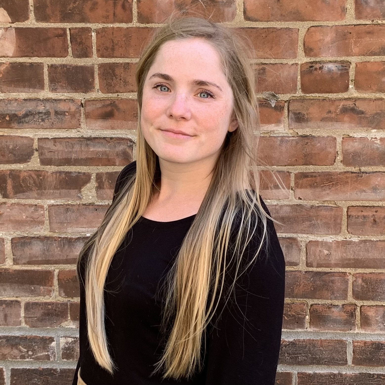Young woman with long blonde hair, wearing a black top, standing in front of a brick wall, looking at the camera with a slight smile.