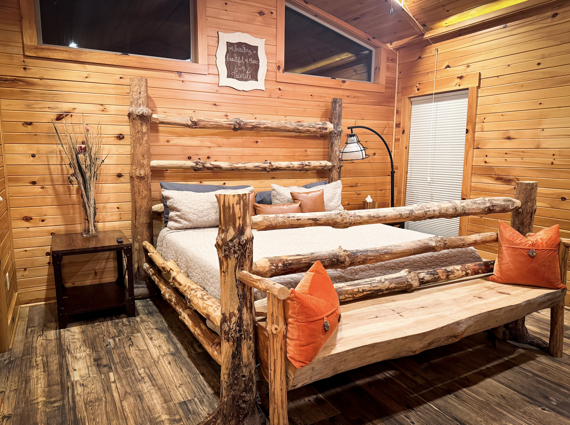 Rustic bedroom with log bed frame, wooden walls, and warm lighting. Decor includes orange pillows, a wall sign, a vase with dried flowers, and a black floor lamp. A window with blinds is on the right wall.