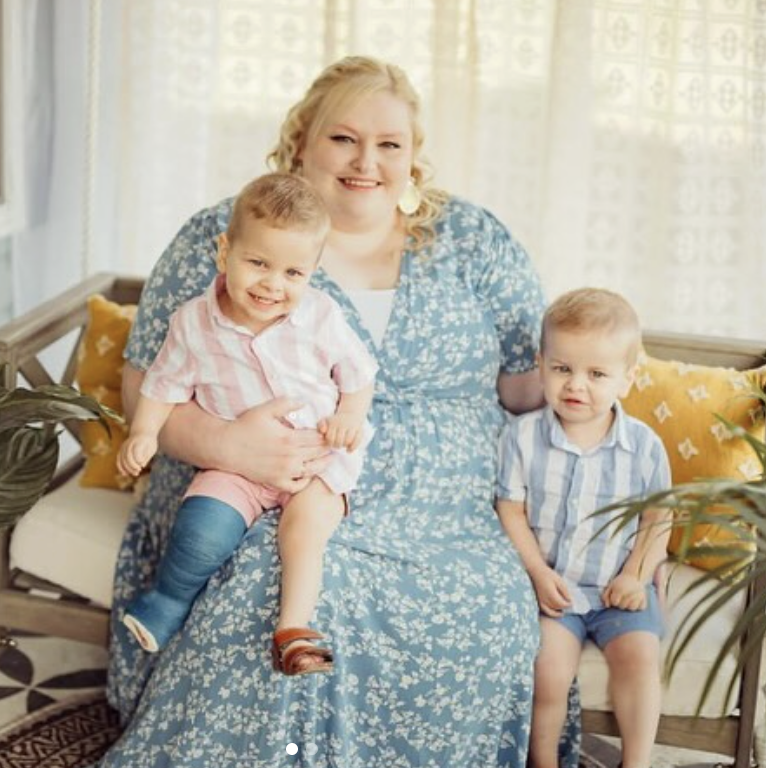 A woman smiling, sitting on a sofa, with two young boys on her lap and beside her, in a brightly lit room with patterned curtains and decorative pillows.