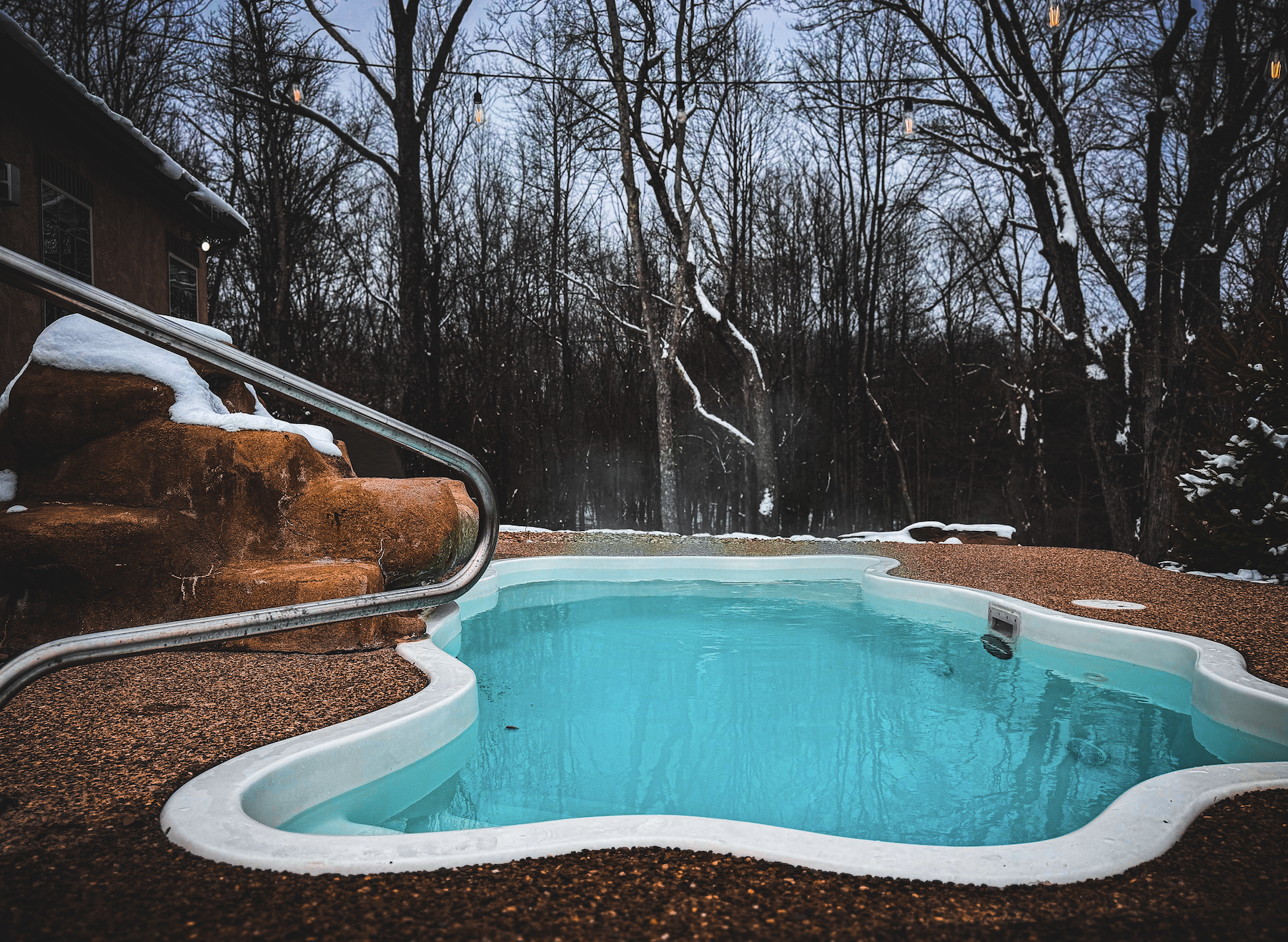 Empty backyard swimming pool with snow on its edges, surrounded by leafless trees and a cloudy sky, in a winter setting.