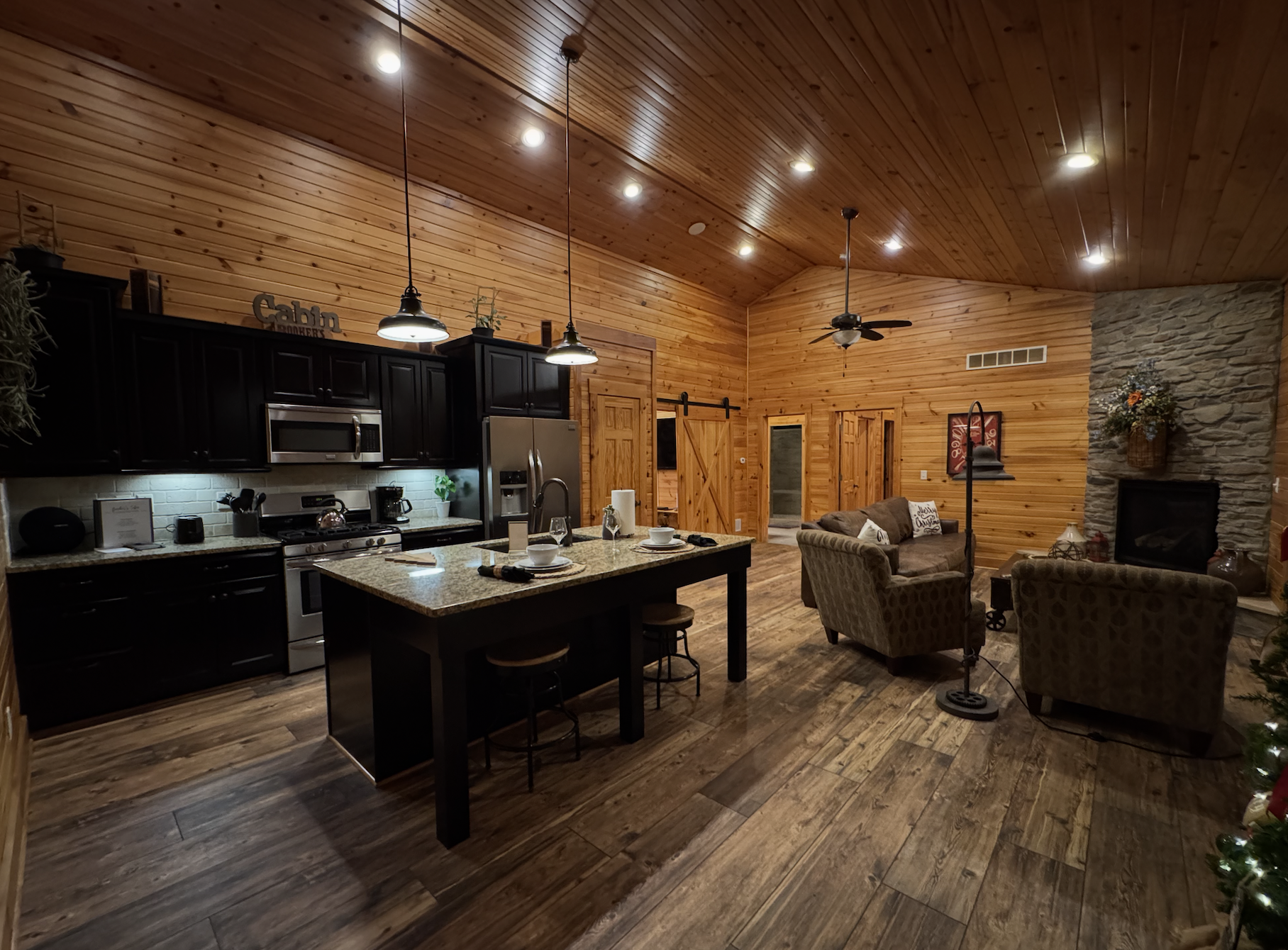 A cozy open-concept living room and kitchen area with wooden walls and ceiling, a stone fireplace, dark kitchen cabinets, a granite island with stools, and seating area with armchairs and a ceiling fan.