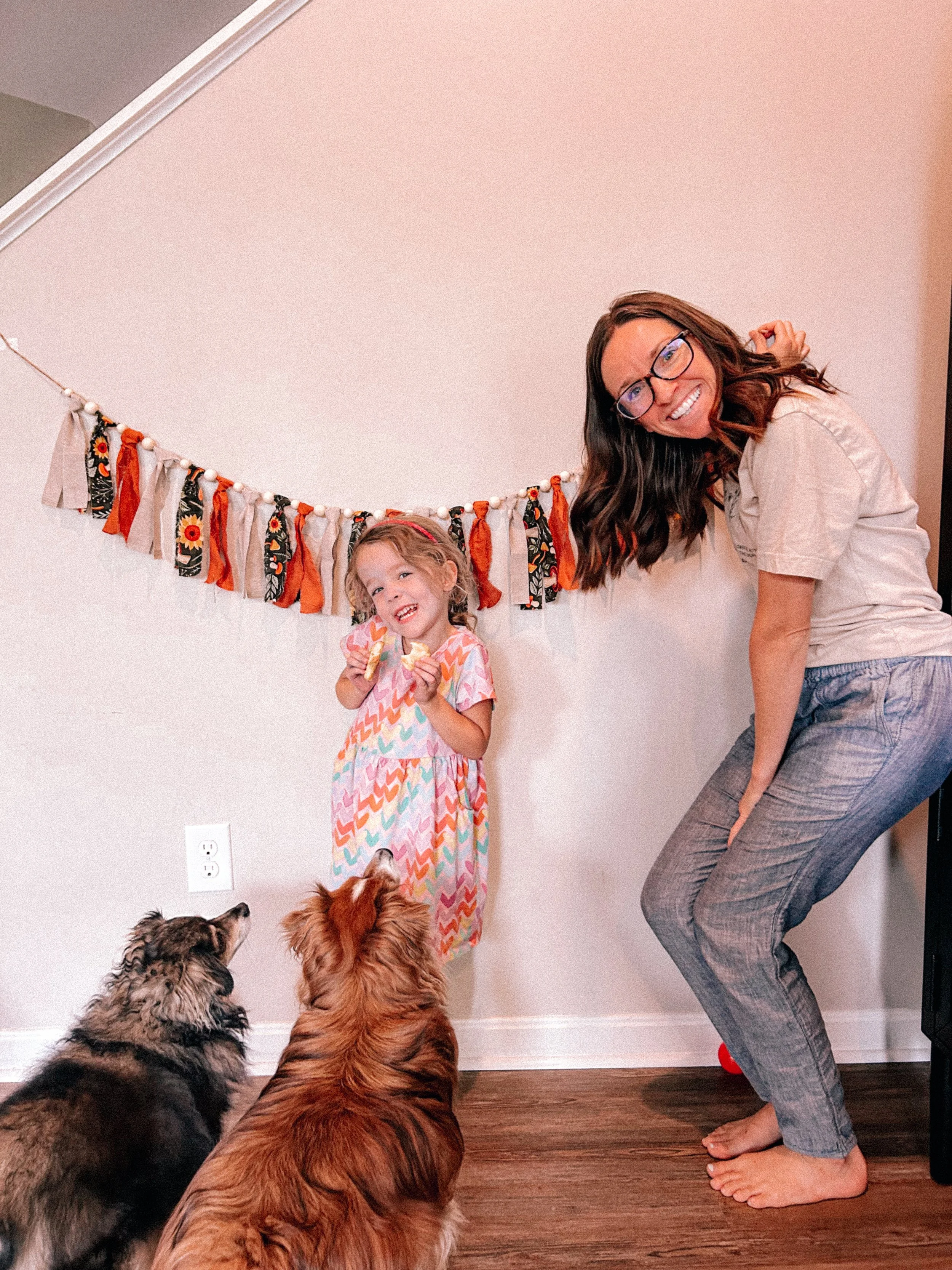 A woman with glasses and long brown hair smiling and dancing with a young girl in a colorful dress, holding treats in her hand, as two dogs watch. They are indoors with a decorative garland hanging on the wall behind them.