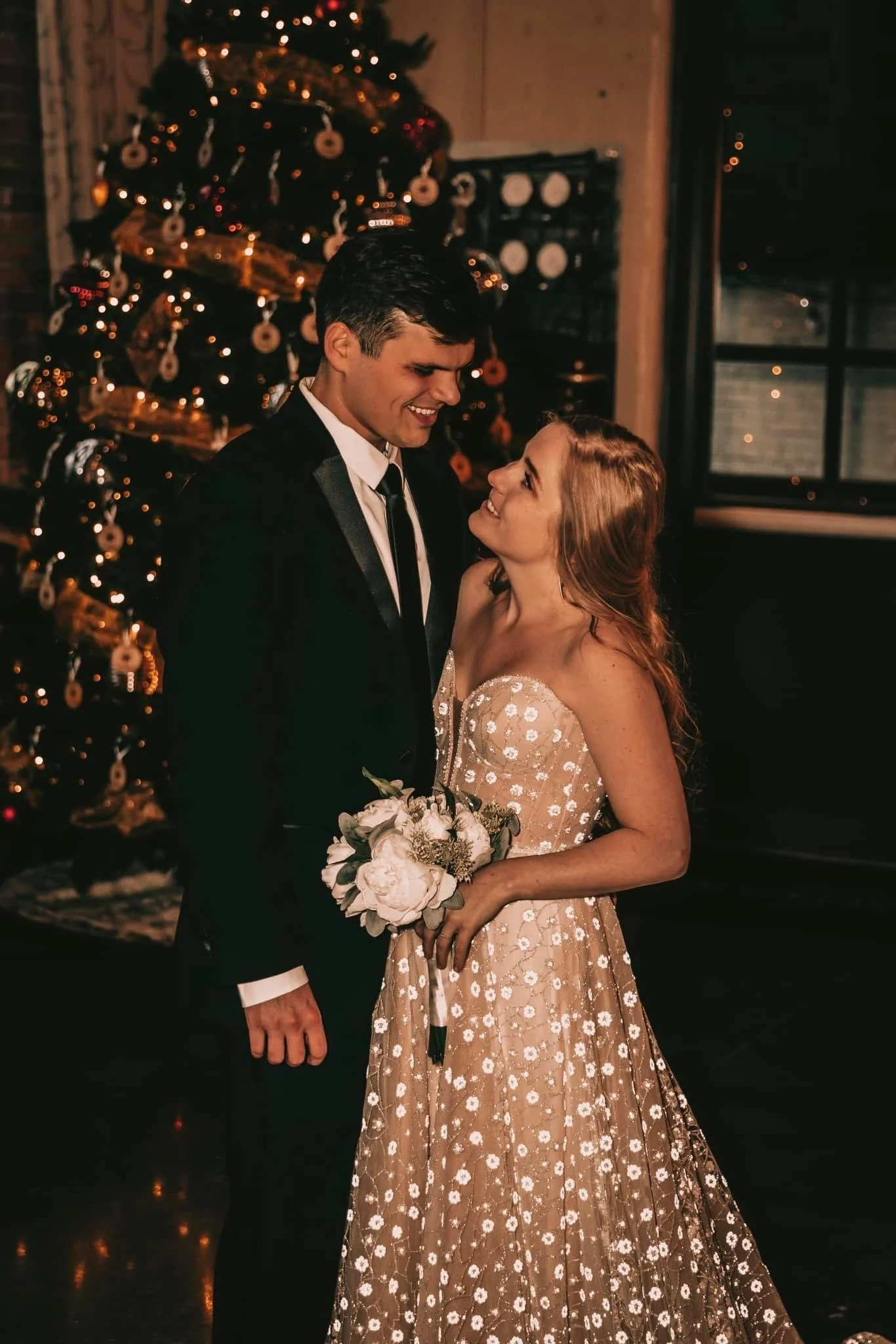 A couple dressed in formal attire smiling at each other in front of a decorated Christmas tree, with the woman holding a bouquet of flowers.