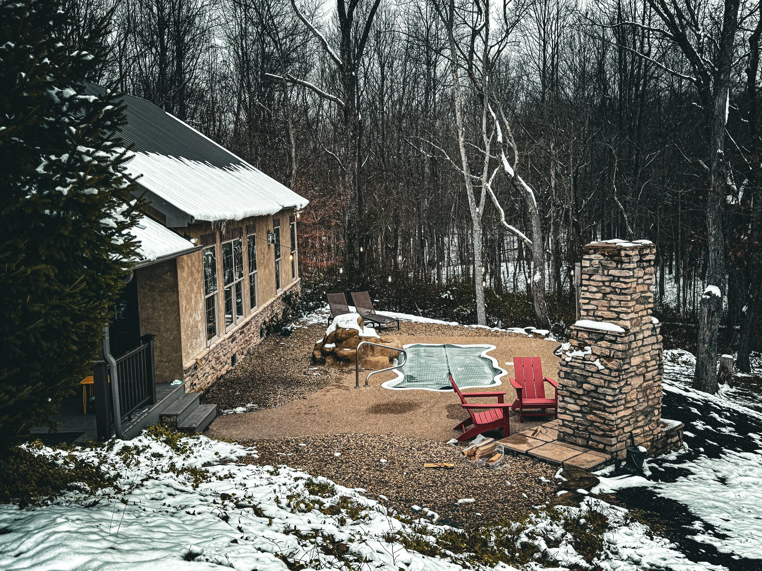A snowy backyard patio with a stone fireplace, red Adirondack chairs, a small swimming pool, and a house with large windows, surrounded by leafless trees.