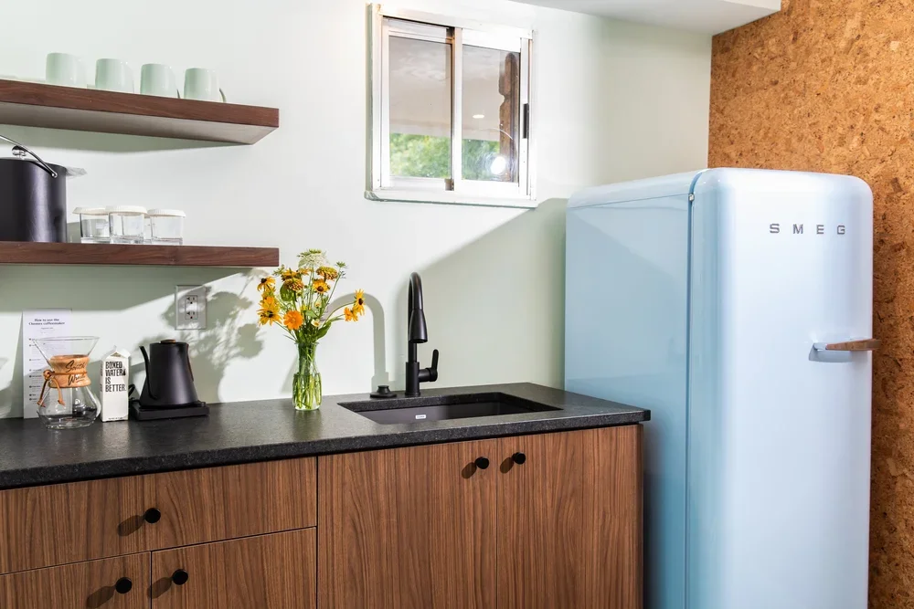 A kitchen corner with a black countertop, a black sink with a black faucet, wooden cabinets, a vase with yellow flowers, open shelves with cups and glasses, a small window, a black kettle, and a light blue vintage-style SMEG refrigerator.
