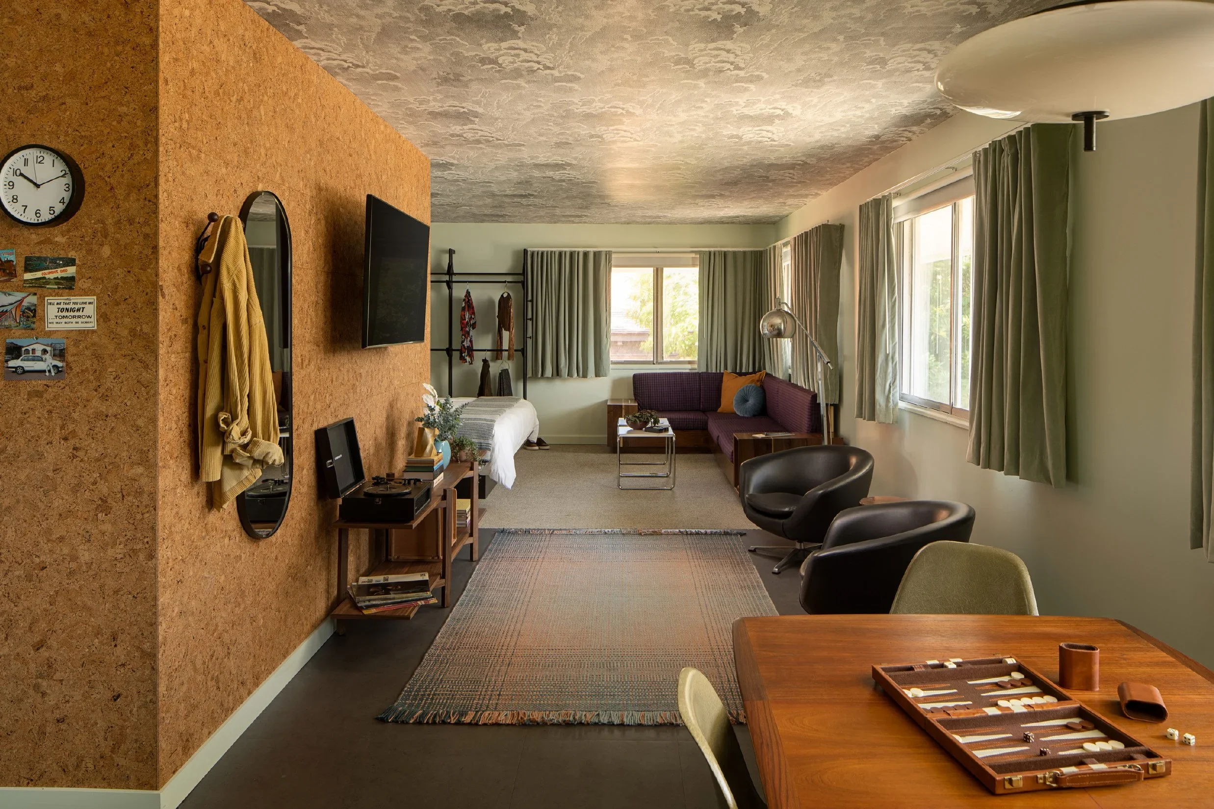 A spacious living room with a cork accent wall, featuring a small tiered wooden side table, a wall-mounted TV, a large window with green drapes, and an L-shaped sofa with cushions. There are two black chairs, a standing lamp, and a wooden dining table with a backgammon set in the foreground. The ceiling is textured, and there is a clock and postcards on the cork wall.
