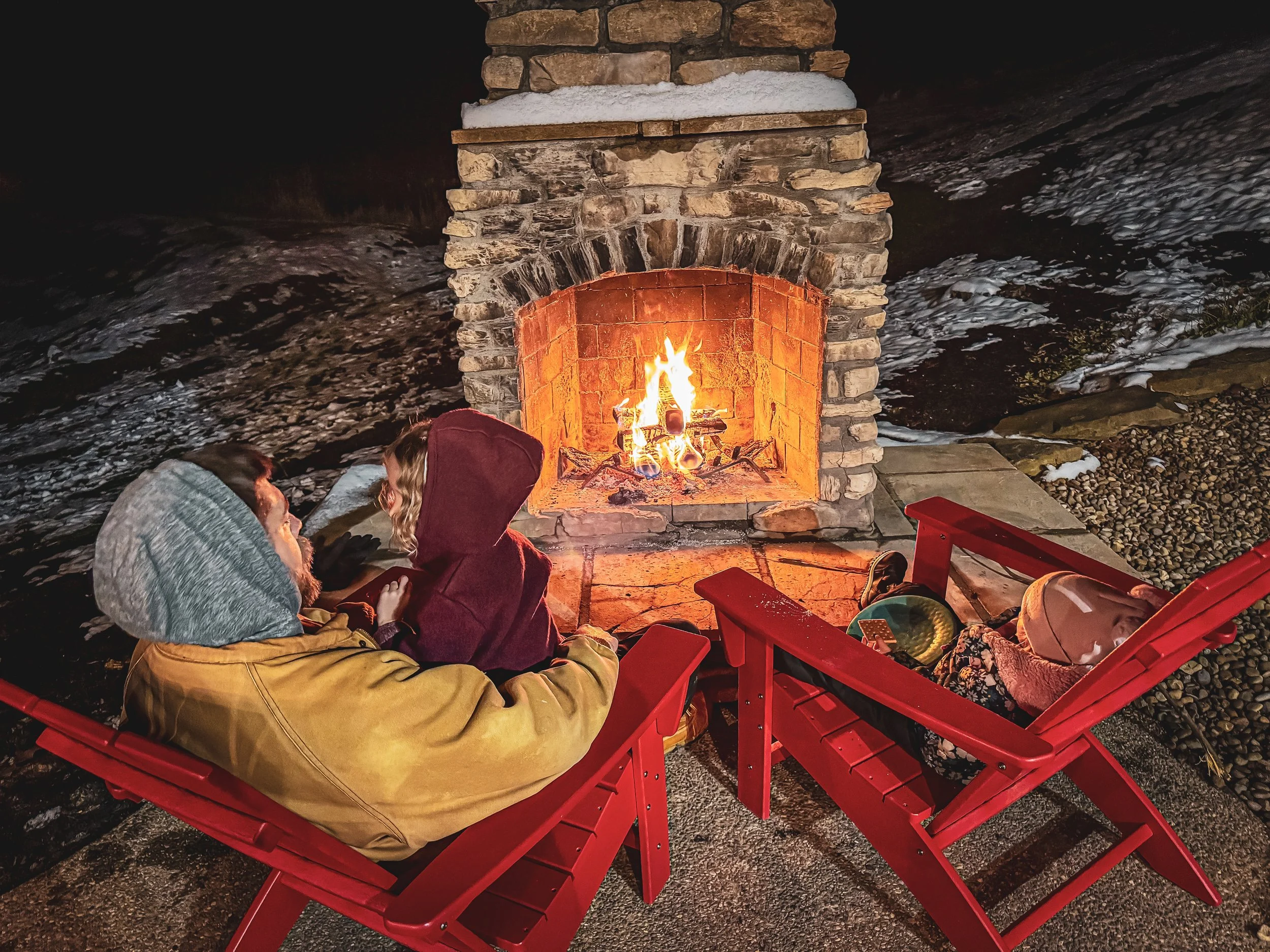 A couple sitting around a campfire at night, surrounded by snow and rocks, on red Adirondack chairs.