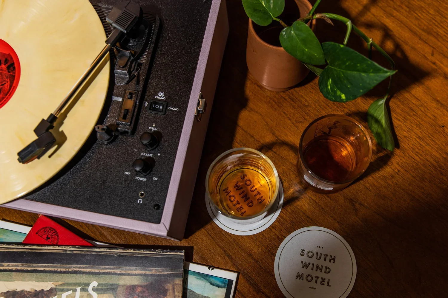 A turntable playing a vinyl record on a wooden table. Two glasses of amber-colored drinks, one with the words "South Wind Motel" underneath a potted plant are on the table. A coaster with the same words is near the glasses.