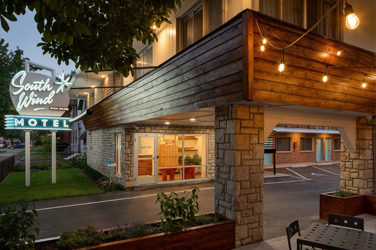 Exterior view of the South Wind Motel at dusk with illuminated signage and string lights on the balcony. The building features brick and wooden elements, with a visible office entrance and parking area.