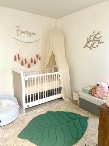 Child's nursery with a white crib, canopy, name 'Emerson' on the wall, pink decorations, a green leaf-shaped rug, and a stuffed pig toy.