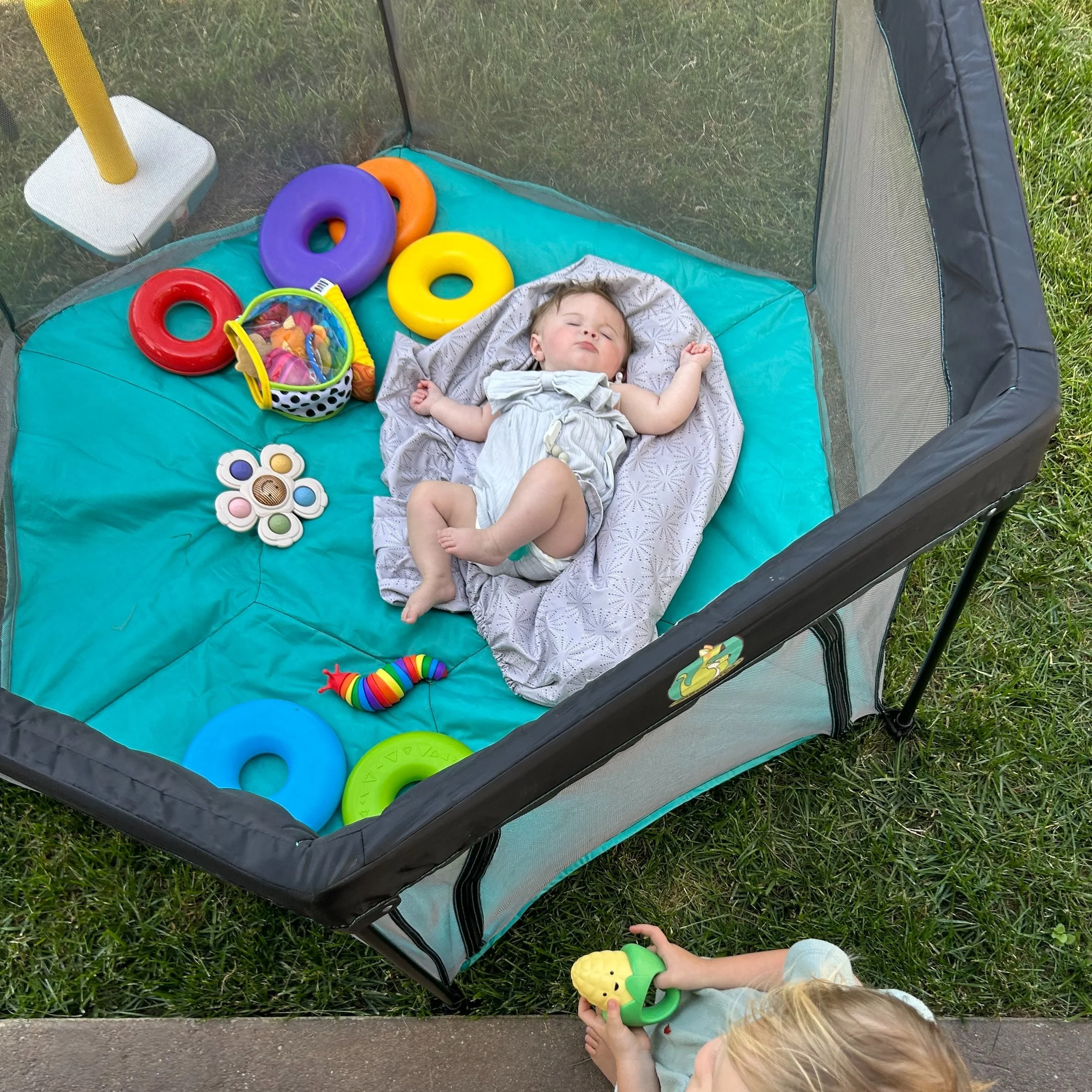 A baby sleeping in a playpen outdoors with colorful plastic rings and toys inside, and a child nearby holding a yellow and green toy.
