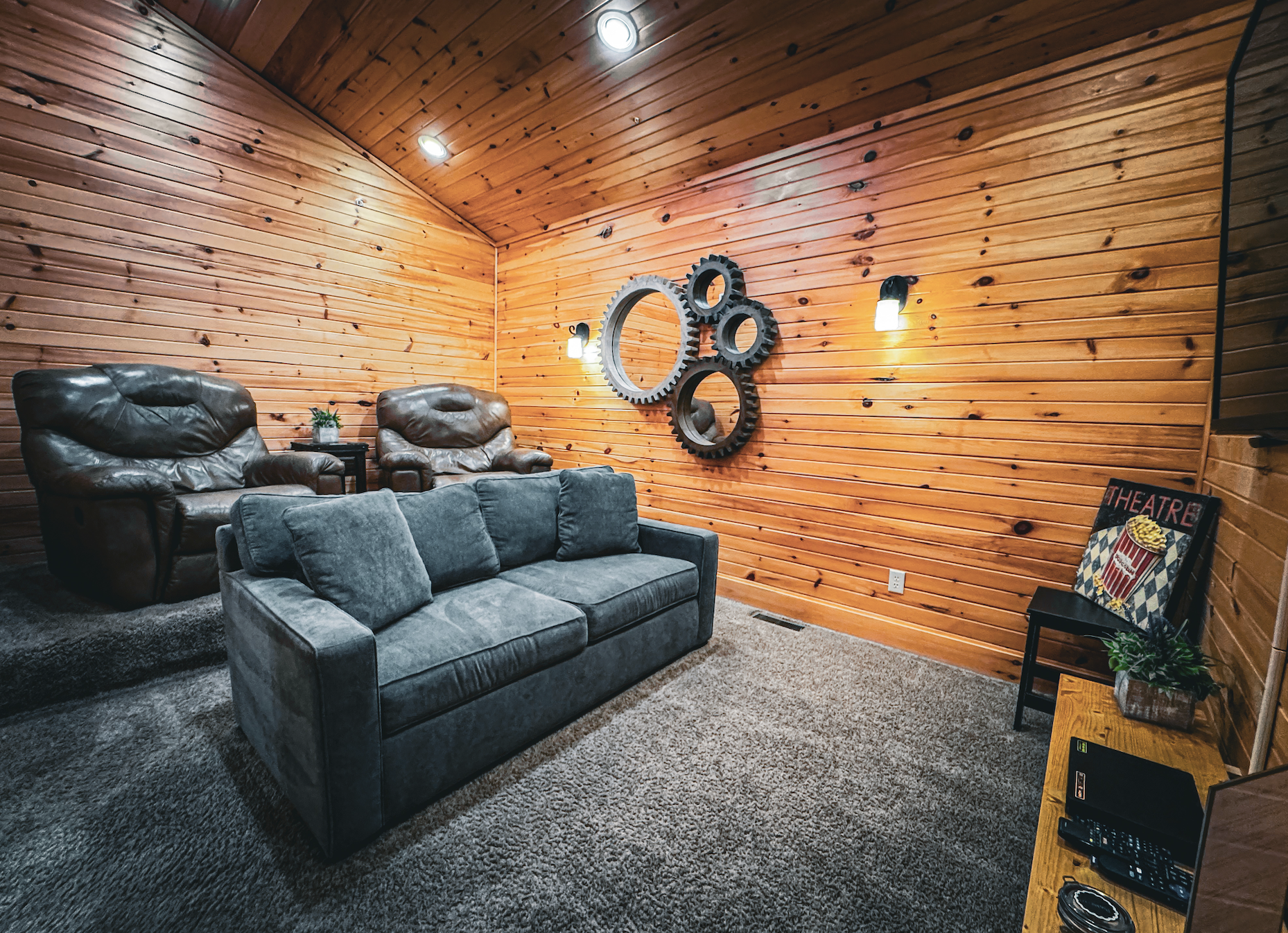 Living room with wooden paneled walls, a gray sofa, two brown leather armchairs, wall-mounted gear-shaped mirrors, wall lights, a small black side table with a plant, a theater poster leaning against the wall, and a wooden cabinet with a potted plant, remote controls, and electronic devices.