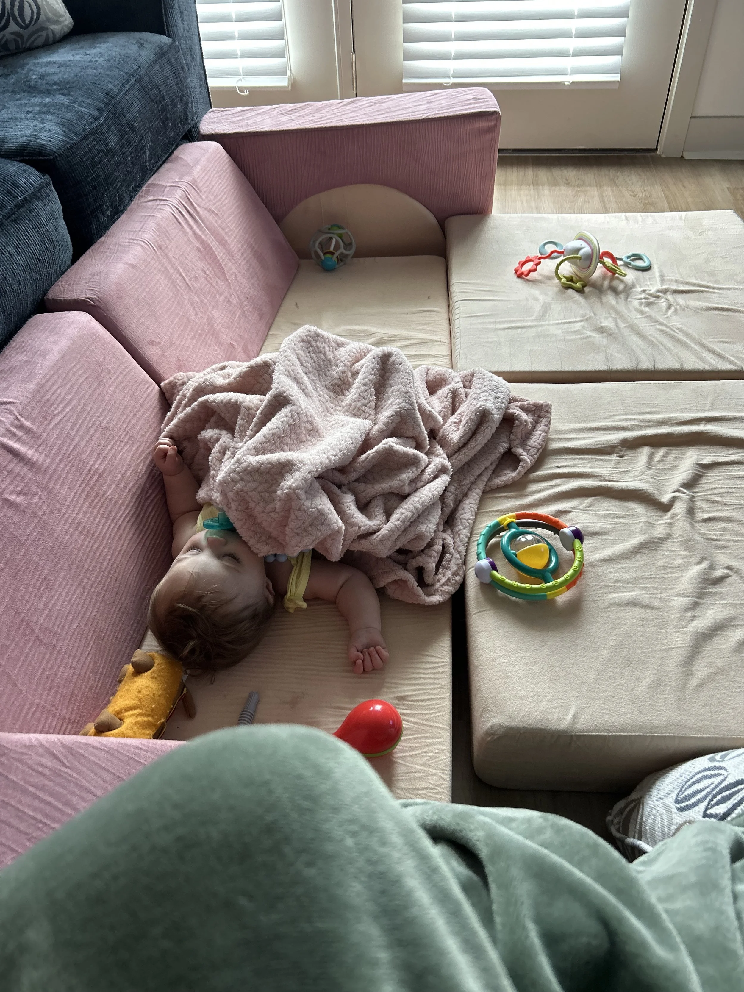 A sleeping baby lying on a Light wood-colored couch with pink cushions, covered with a pink blanket. The baby is holding a toy giraffe and has a pacifier. There are colorful toys on the couch and the adjoining cushions, with a glass door with blinds in the background, letting in natural light.